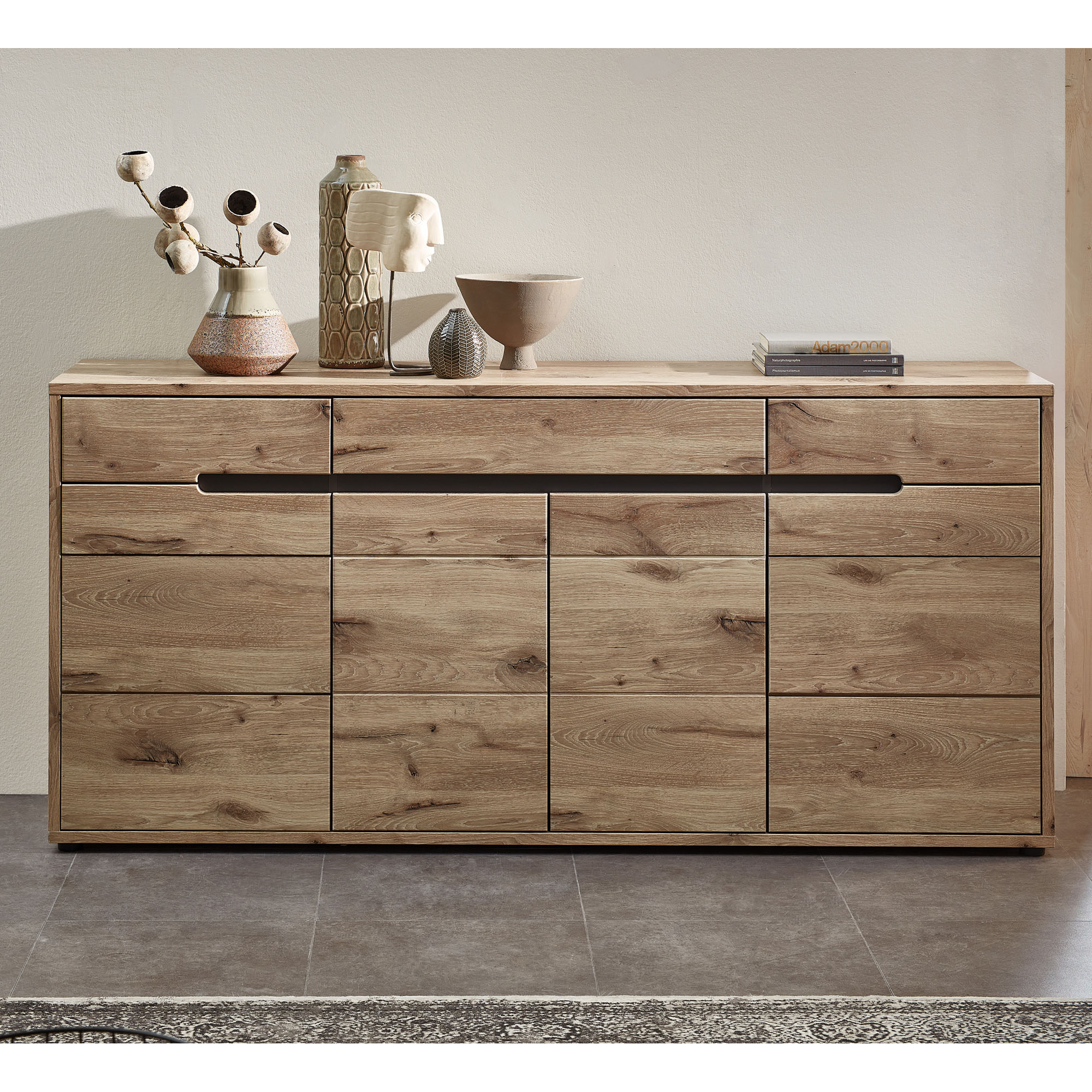 Das Sideboard steht vor einer hellen Wand auf einem Fliesenboden. Oben darauf befinden sich mehrere Dekoobjekte und ein Bücherstapel.