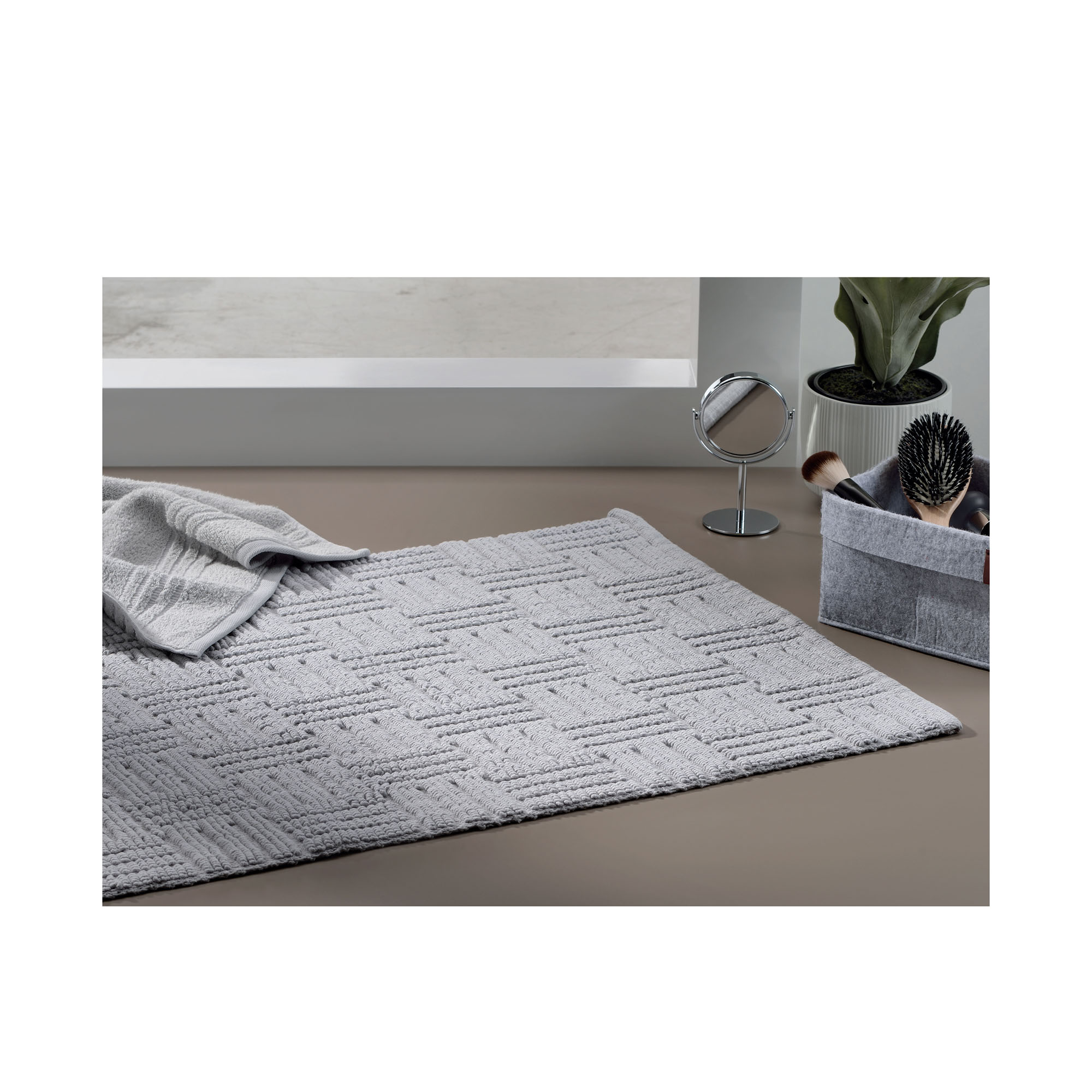 Weiße Badematte Leana mit geometrischer Struktur auf hellem Boden neben Korb, Handtuch und Pflanze.