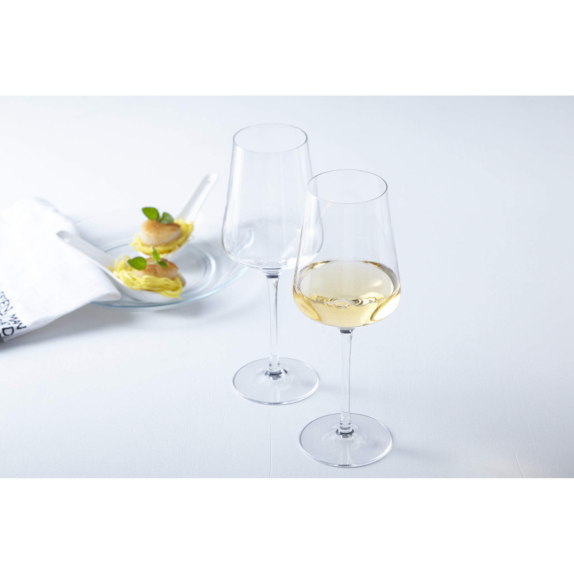 Zwei Weißweingläser eines gefüllt mit Weißwein auf hellem Tisch mit Dessert