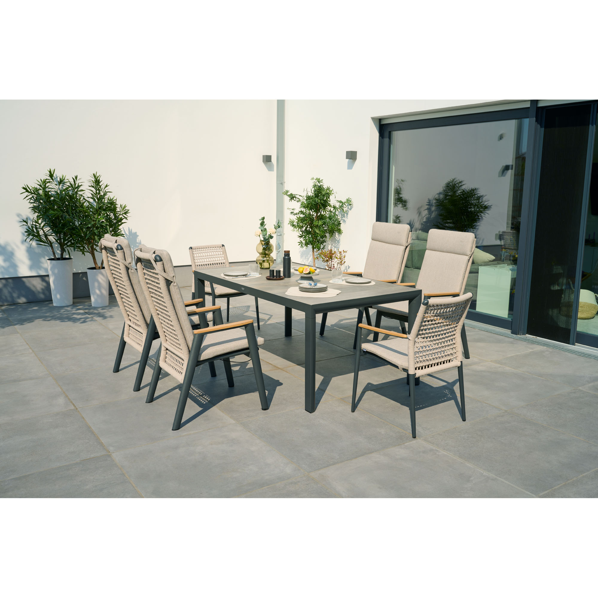 Mehrere Stühle mit beige-grauen Polstern stehen um einen großen rechteckigen Tisch auf einer Terrasse, im Hintergrund Pflanzen und eine Glasfassade.