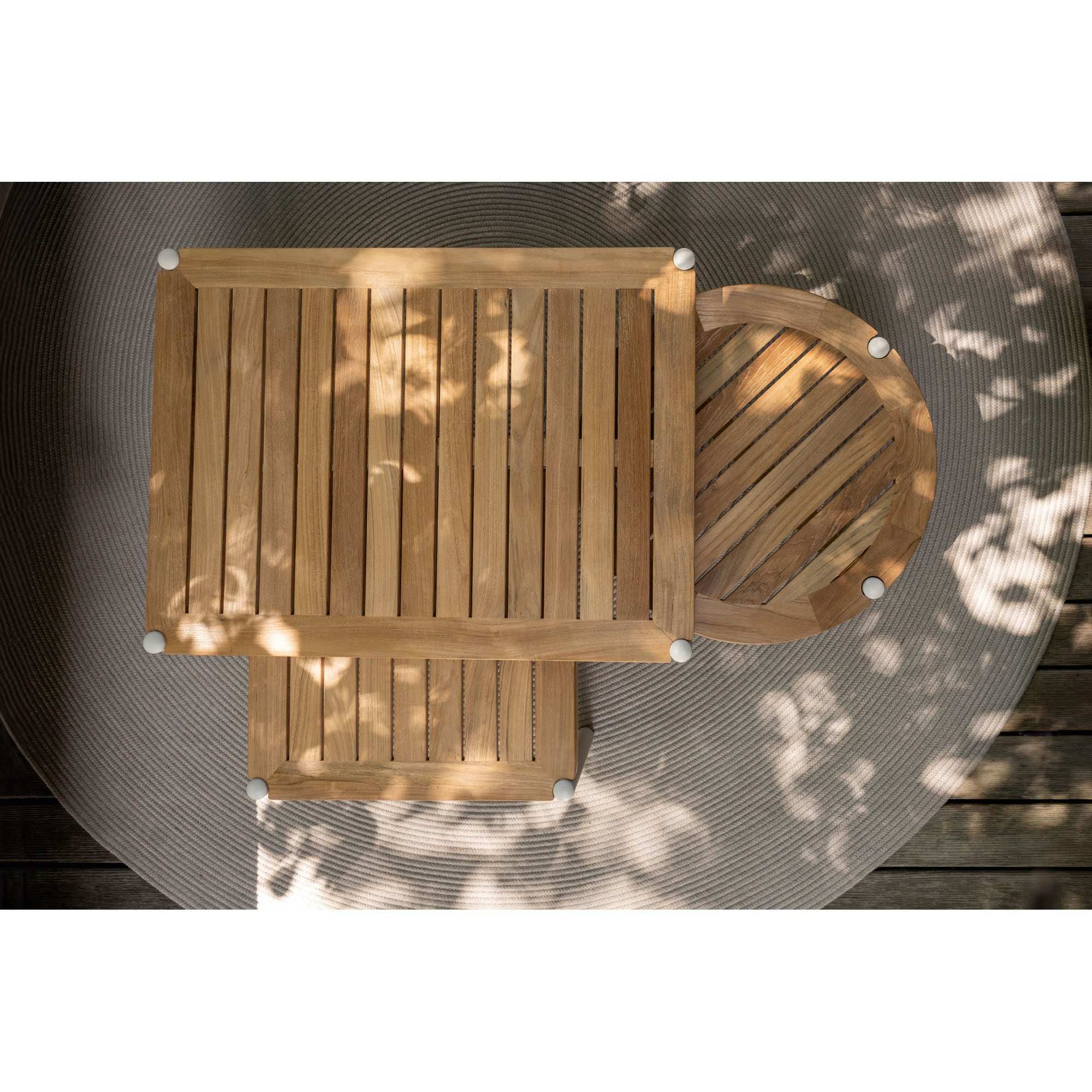 Draufsicht auf zwei rechteckige und einen runden Tisch mit Holzlatten-Oberflächen, teilweise von Blättern beschattet, auf einem runden Teppich arrangiert.