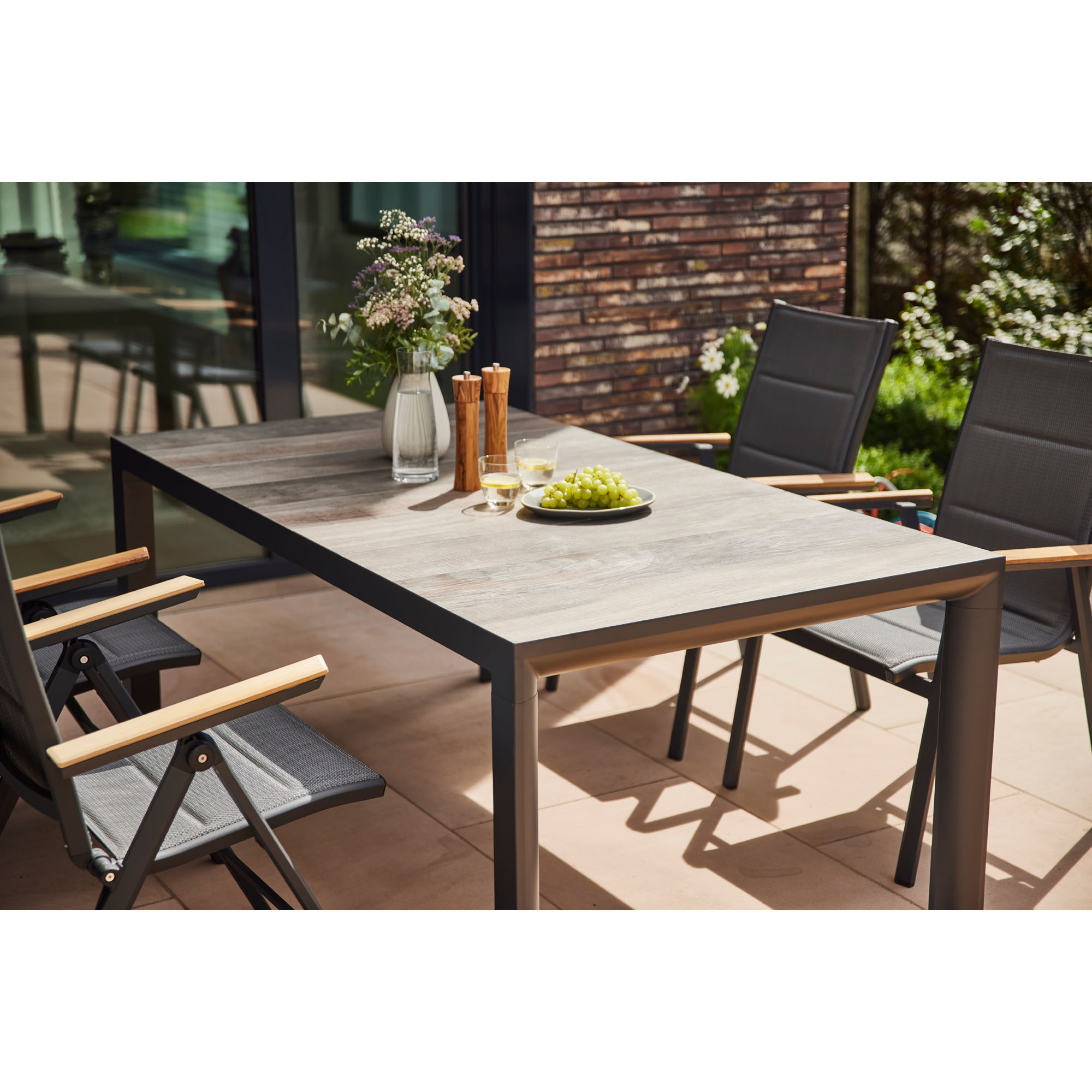 Gartentisch mit dunkelgrauem Gestell und Holzdekor-Tischplatte, umgeben von Gartenstühlen, gedeckt mit Gläsern, Blumen und einem Teller Trauben auf einer sonnigen Terrasse.