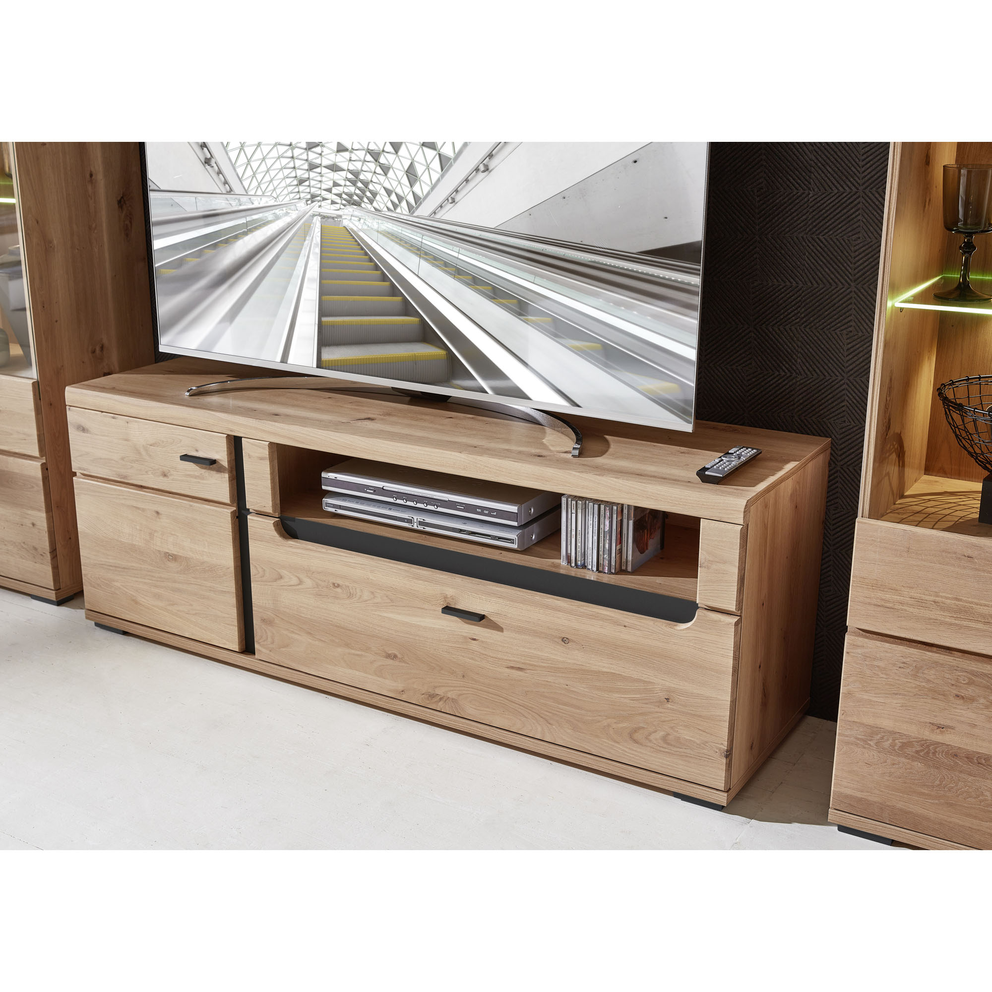 Seitliche Ansicht eines TV-Lowboards aus hellem Holz mit Fernseher; offenes Technikfach mit Geräten und CDs sowie geschlossene Fächer sind sichtbar.