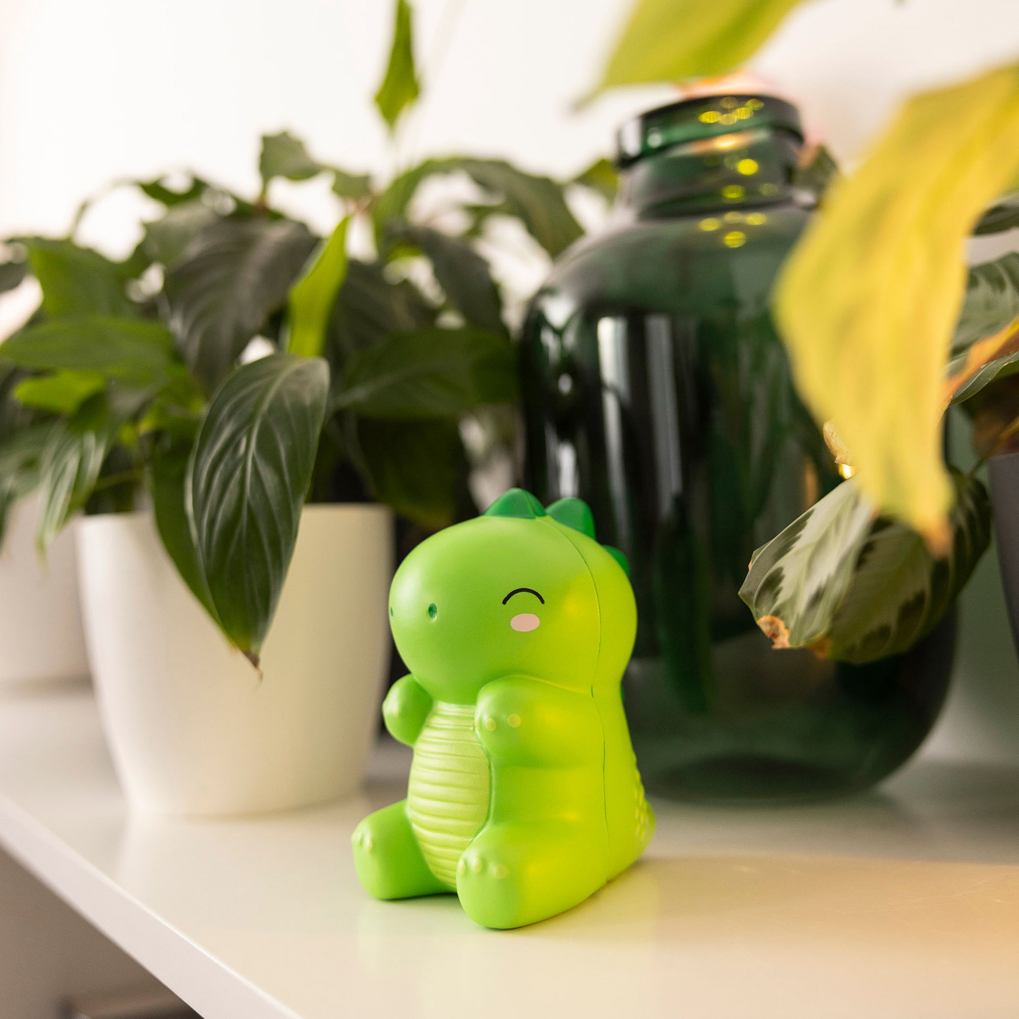 Grüne Antistress-Figur in Dinosaurierform, auf einer hellen Fläche vor mehreren Zimmerpflanzen dargestellt.