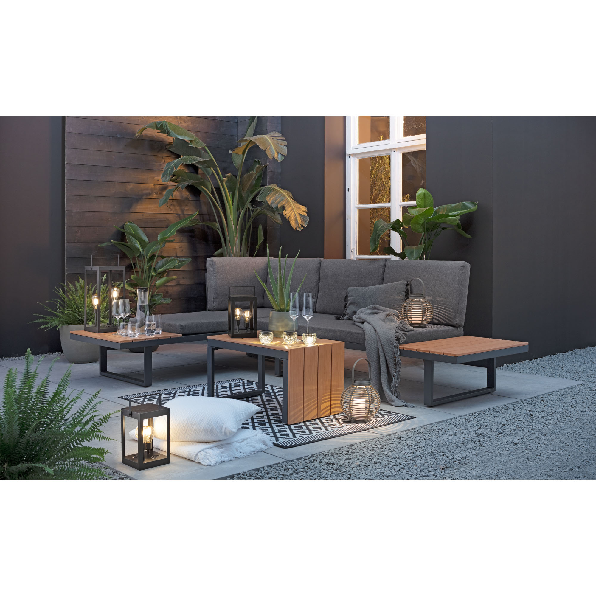 Gartensofa mit Polstern und niedriger Tisch auf einer Terrasse mit Pflanzen und Beleuchtung im Hintergrund