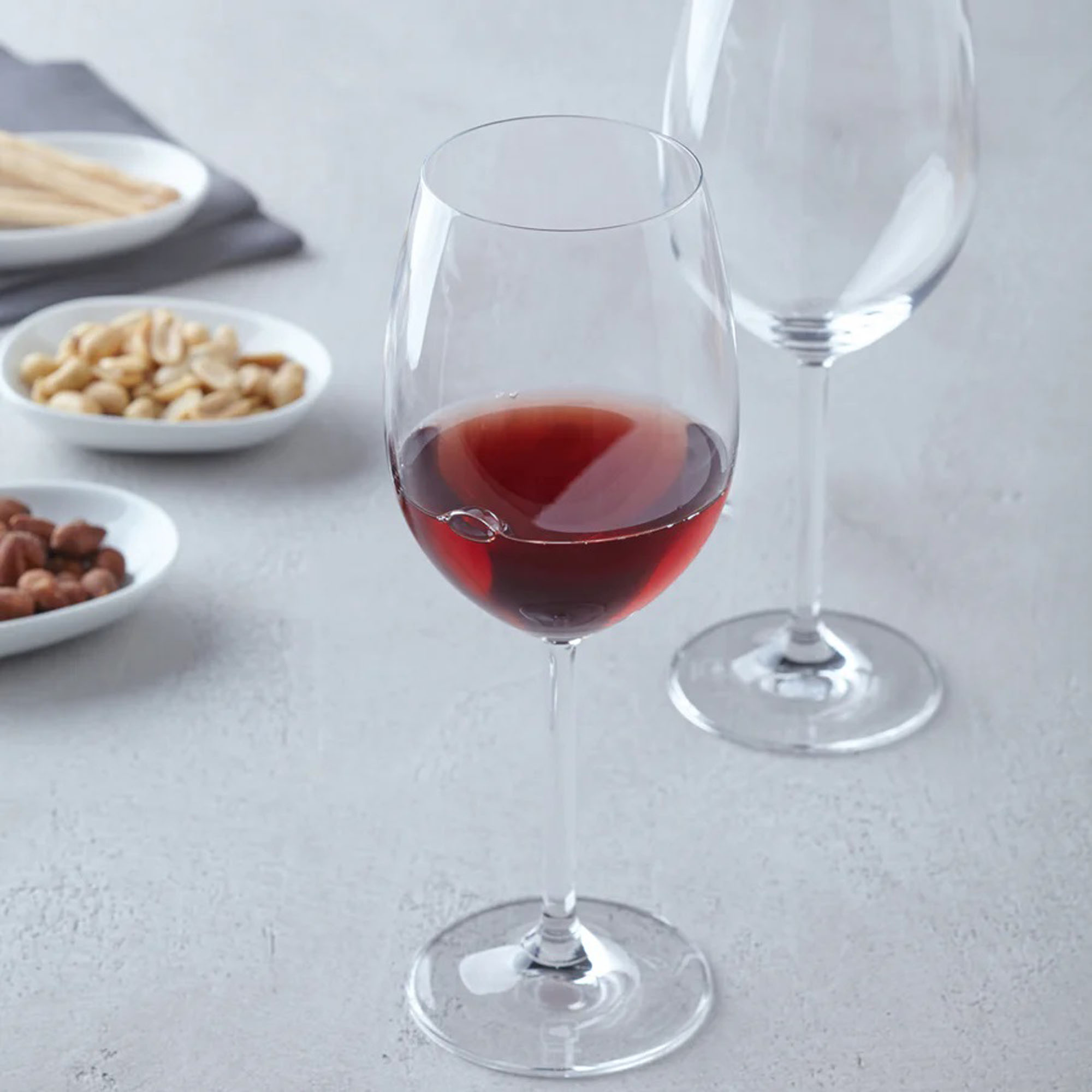 Rotweinglas mit rotem Wein auf hellem Tisch daneben Snacks wie Nüsse und ein weiteres leeres Weinglas im Hintergrund