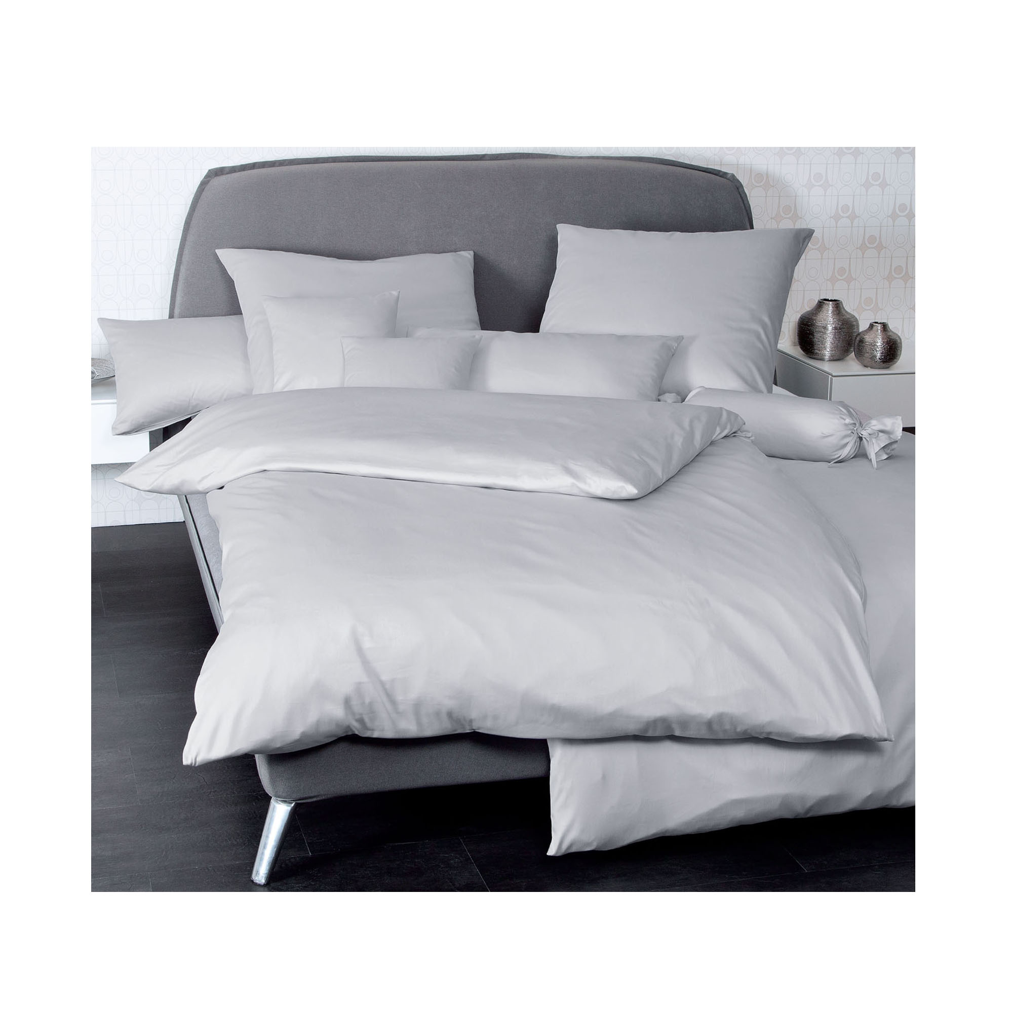 Bett mit Janine Mako-Satin Bettwäsche Colors 31001 in Platin, arrangiert mit mehreren Kissen in einem hellen Schlafzimmer.