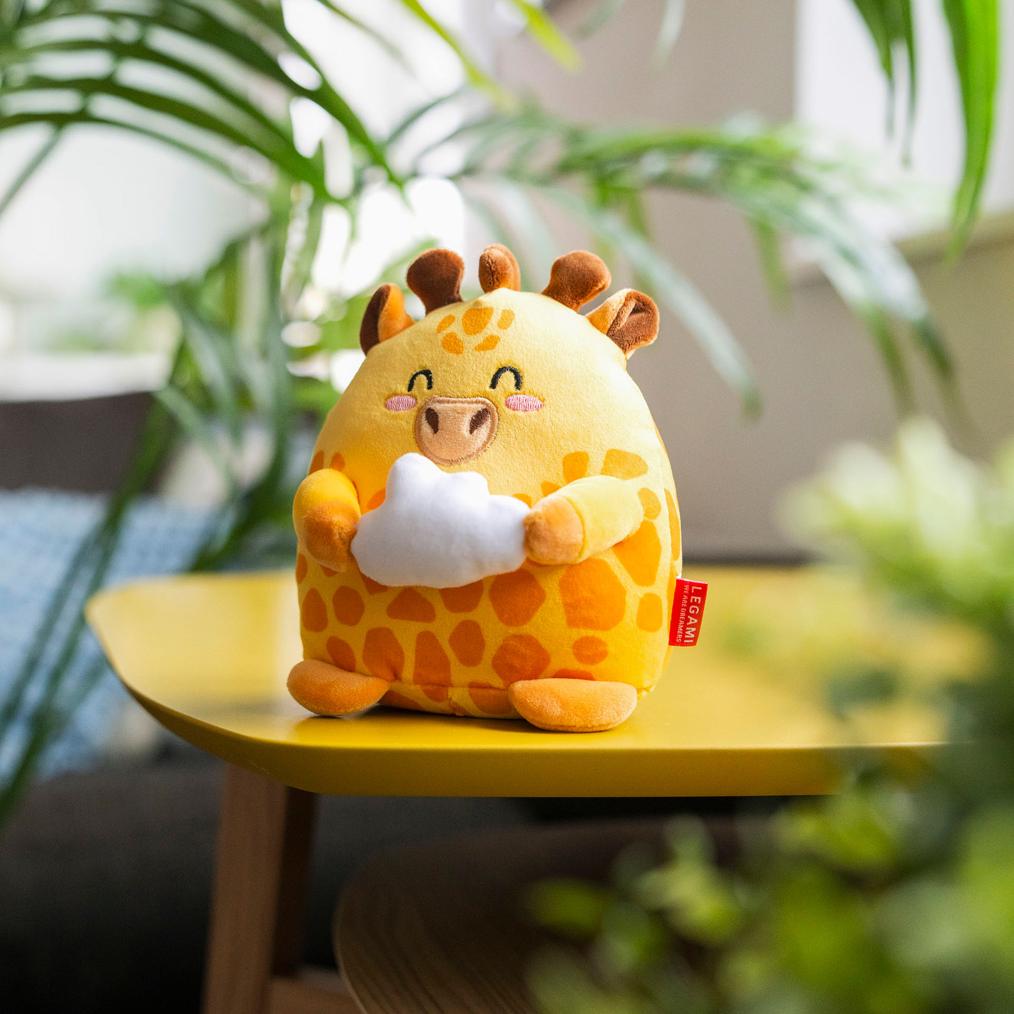 Orangefarbenes Giraffen-Plüschtier, auf einem gelben Beistelltisch zwischen Pflanzen dargestellt.