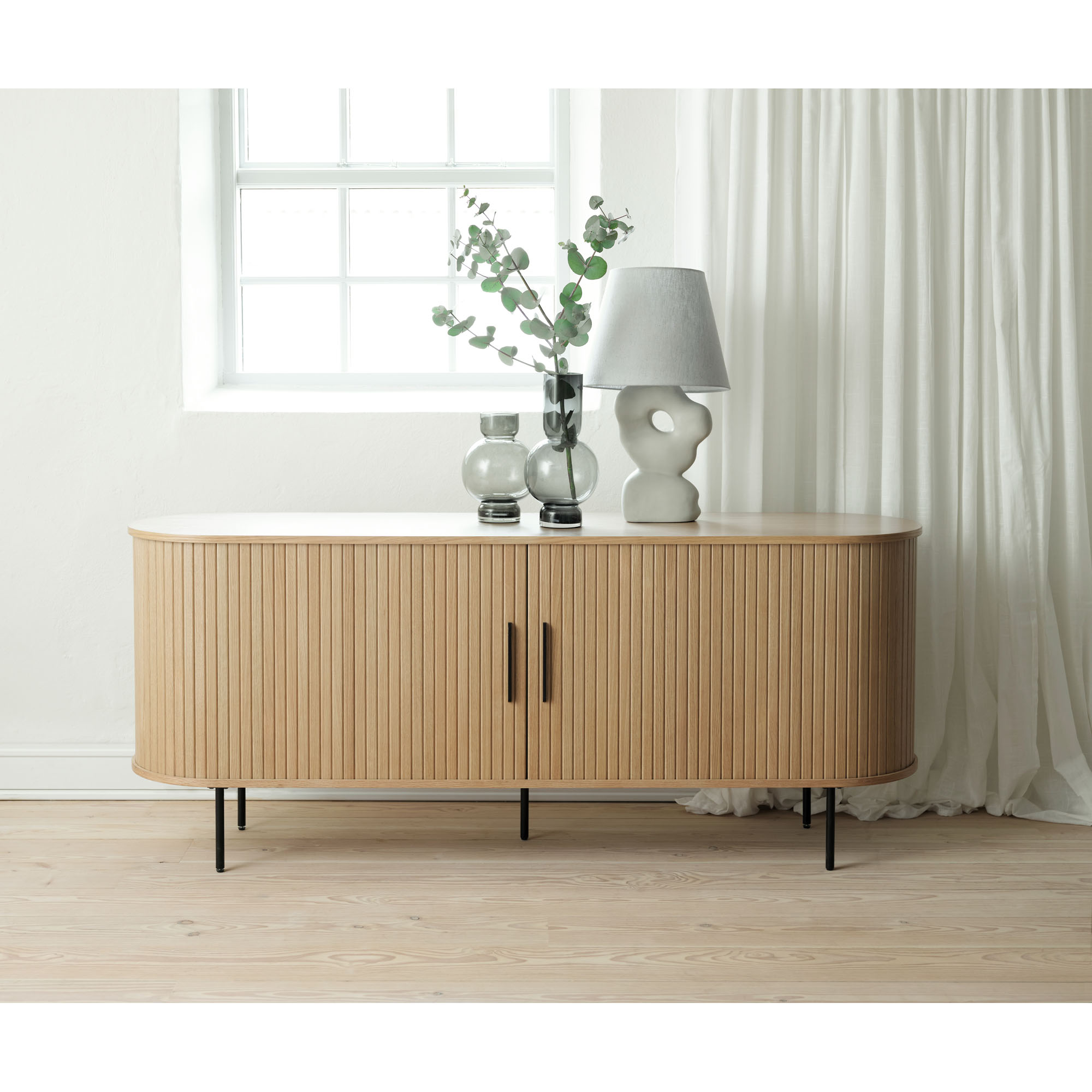 Modernes Sideboard mit Lamellenfront in Holzoptik, dekoriert mit Vasen und Tischlampe, vor hellem Fenster und weißen Vorhängen.