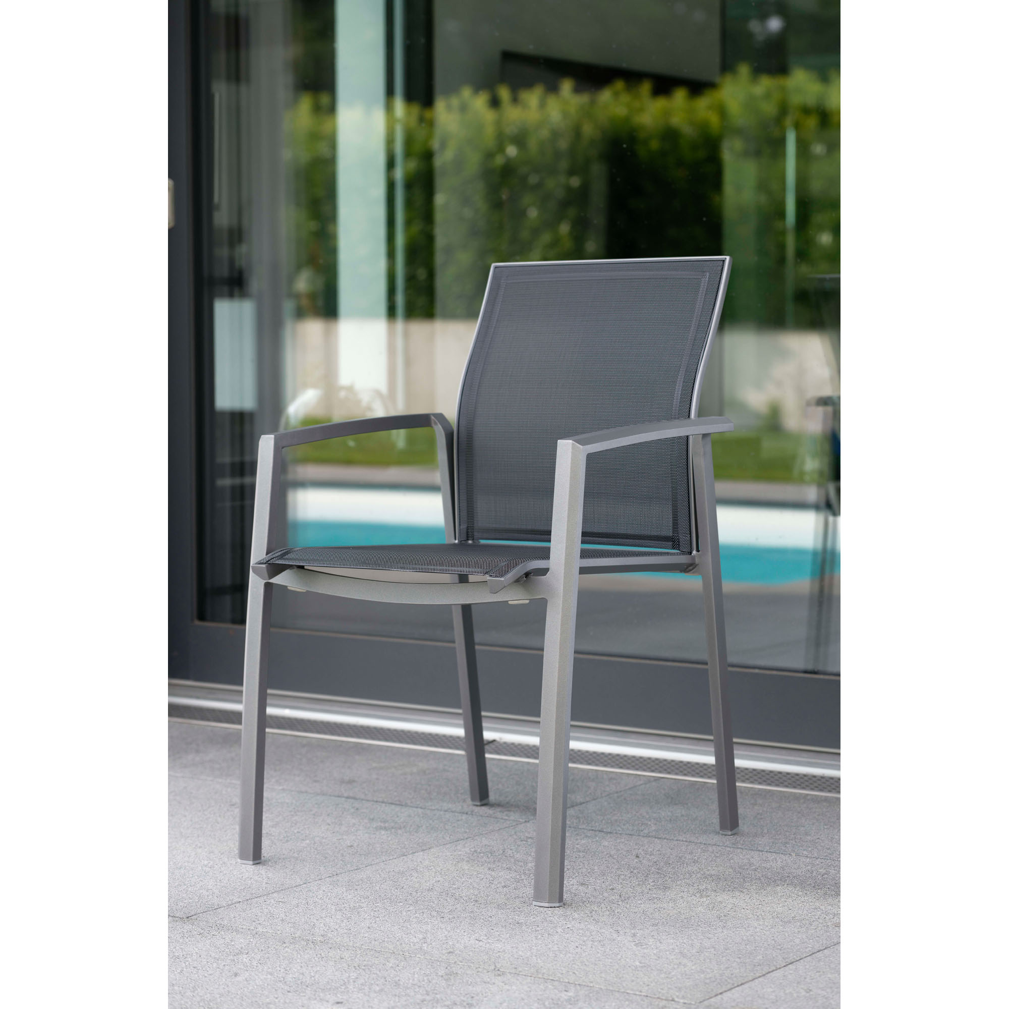 Ein grauer Stuhl mit Netzbespannung steht auf einer Terrasse vor einer Glasfront mit Blick auf einen Garten und einen Pool.