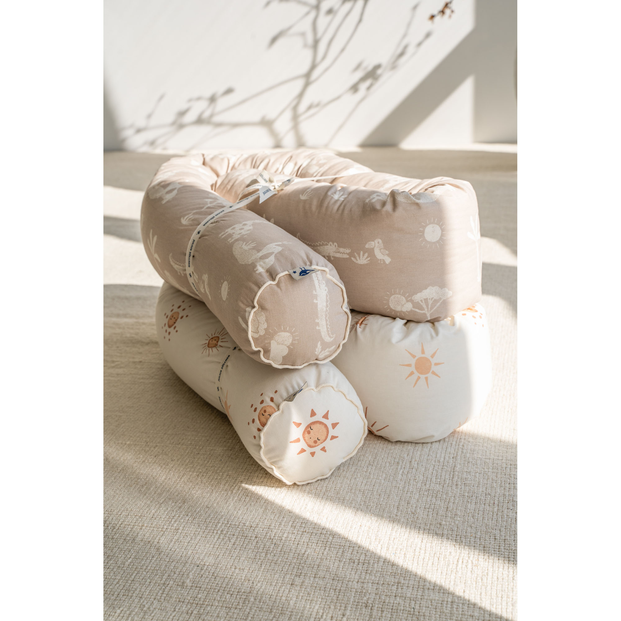 Zwei Baby Bettschlangen aufeinandergelegt, eine mit Sonnendruck in Beige, die andere mit Safari-Muster, auf hellem Teppich im Sonnenlicht