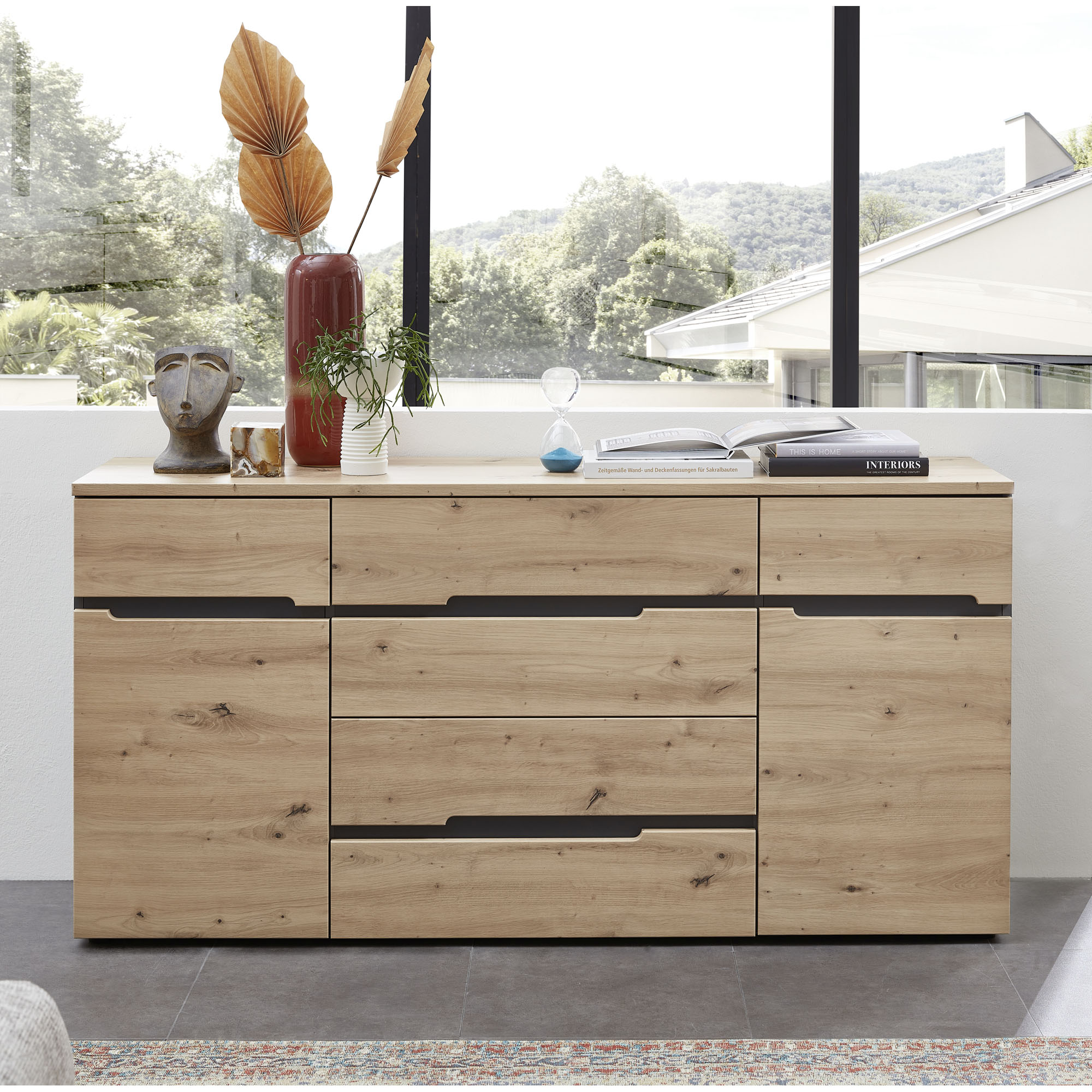 Holzsideboard vor großer Fensterfront; darauf Vasen, eine Pflanze, eine Sanduhr und ein geöffnetes Buch.