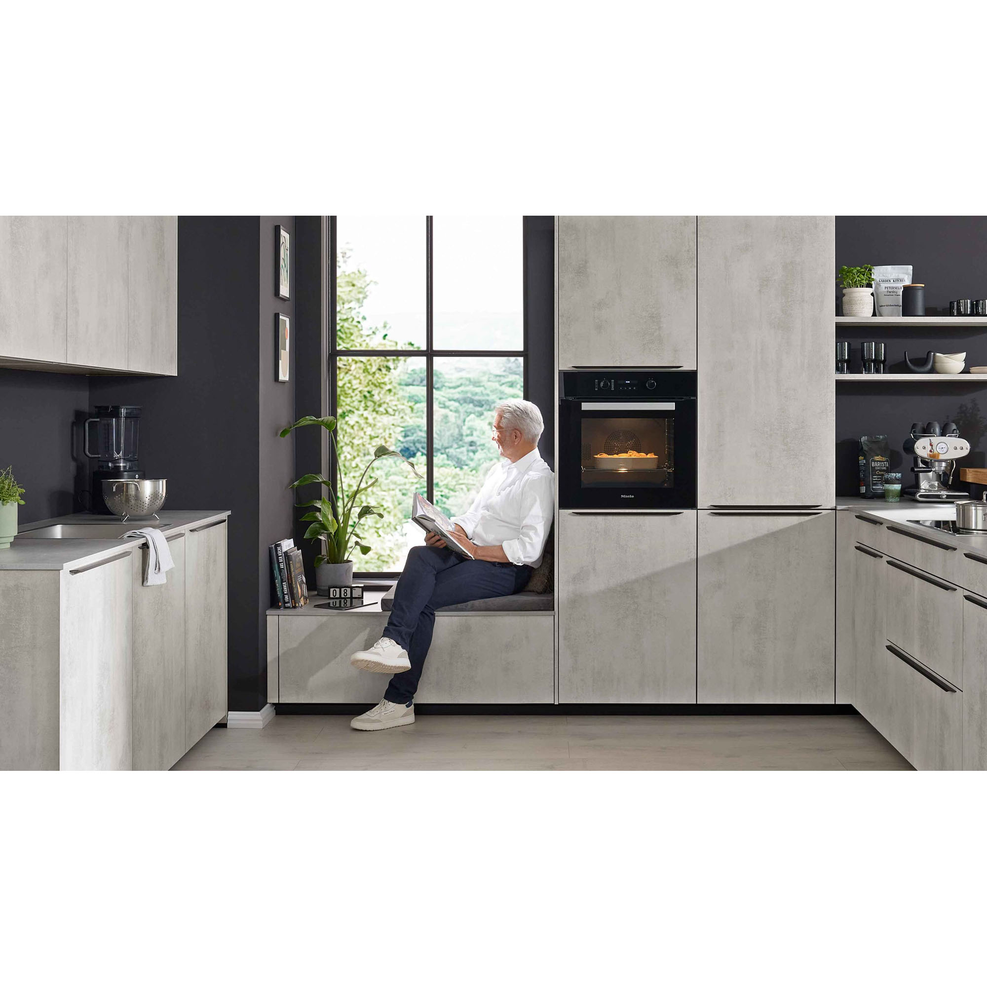 Person sitzt auf einer Fensterbank in einer modernen Küche mit grauen Fronten und integriertem Backofen
