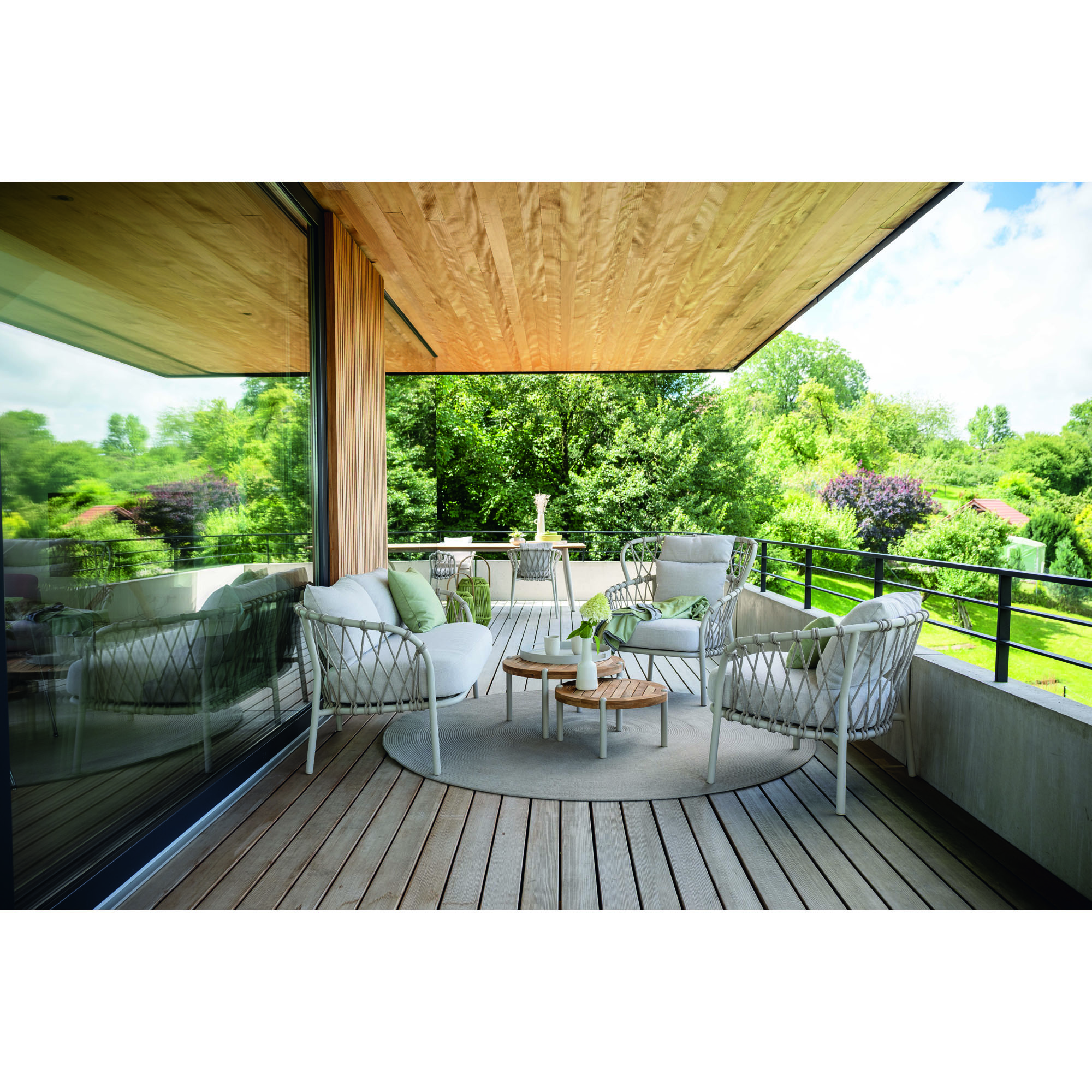 Terrassenszene mit mehreren Sesseln und zwei runden Beistelltischen aus Holz vor einer Glasfassade und Blick ins Grüne