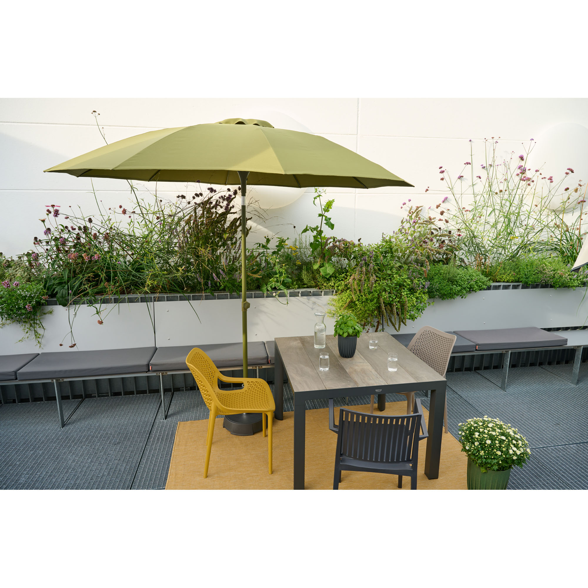 Ein quadratischer Tisch mit Stühlen steht auf einer Terrasse, darüber ein grüner Sonnenschirm. Im Hintergrund sind Pflanzen und Sitzbänke.