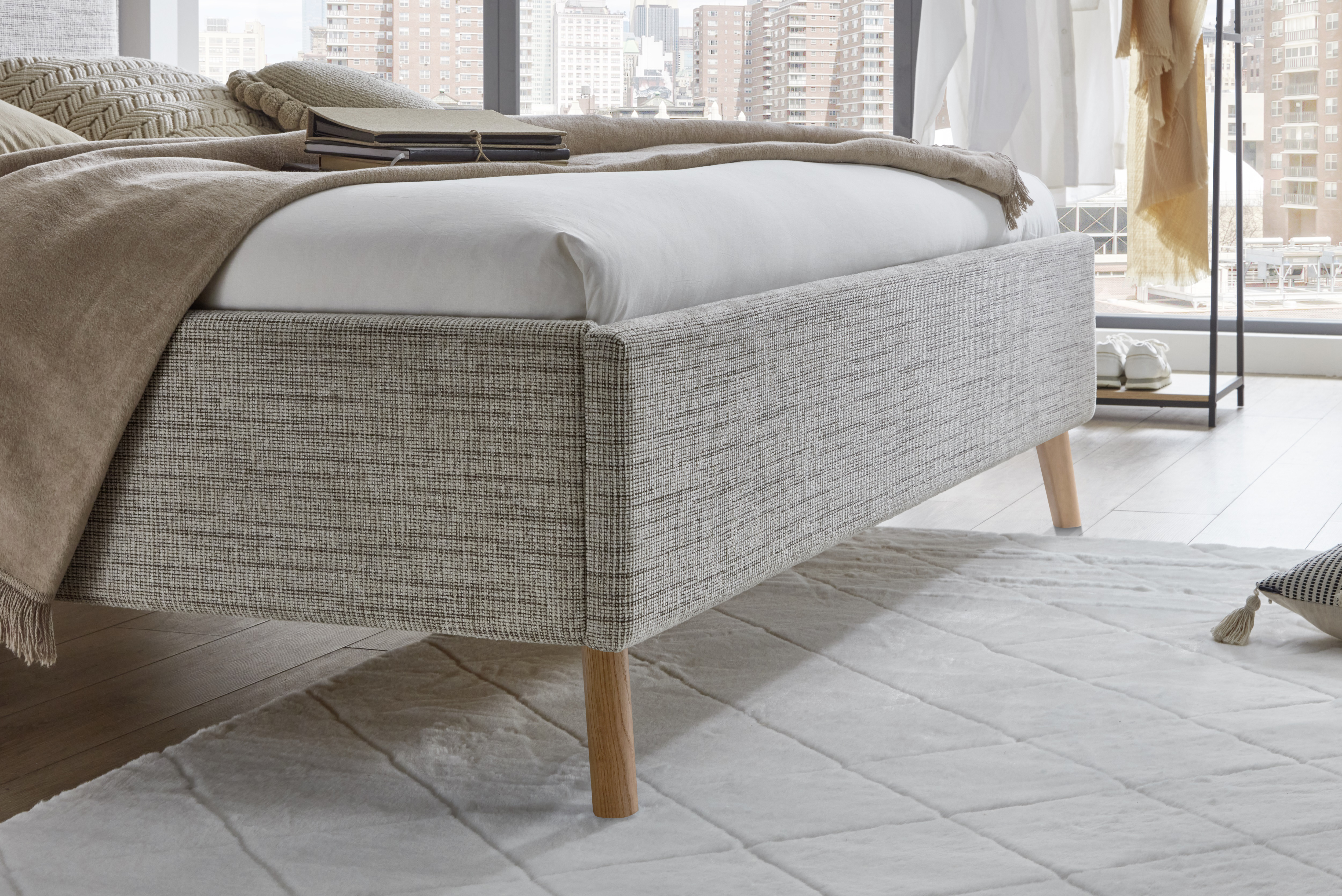 Nahaufnahme eines hellen Polsterbetts mit Holzfüßen, strukturierter Stoffbezug, weißer Bettdecke und brauner Tagesdecke in modernem Schlafzimmer