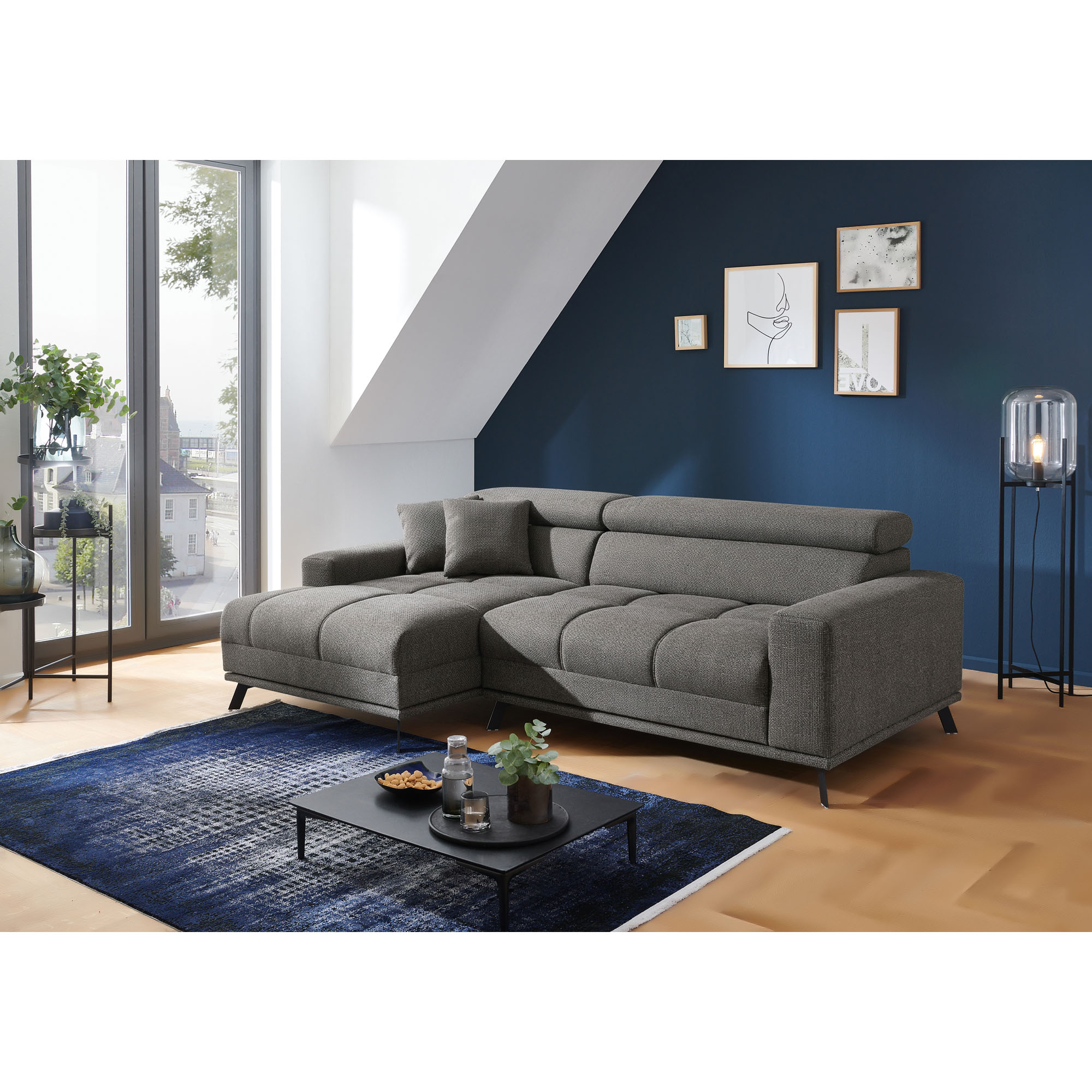 Dunkelgraue Eckcouch mit Longchair links im Bild, zwei Kissen und verstellbaren Kopfstützen vor einer dunkelblauen Wand