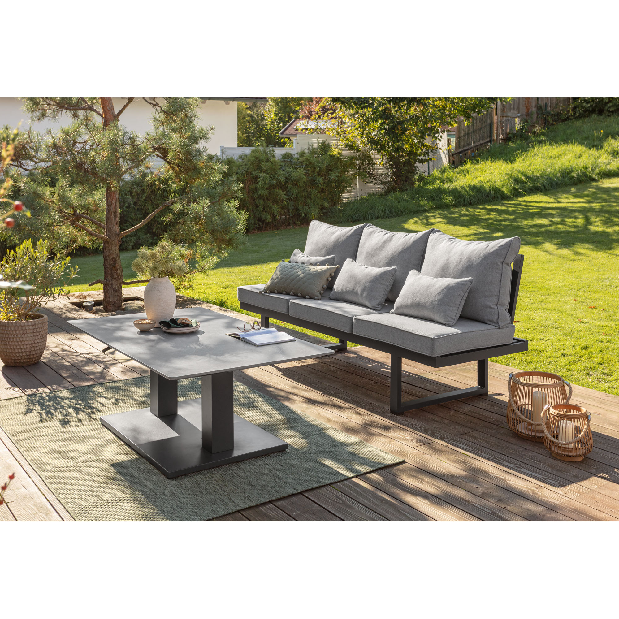 Gepolsterte Gartenbank neben einem rechteckigen Tisch auf einer Holzterrasse mit Garten im Hintergrund
