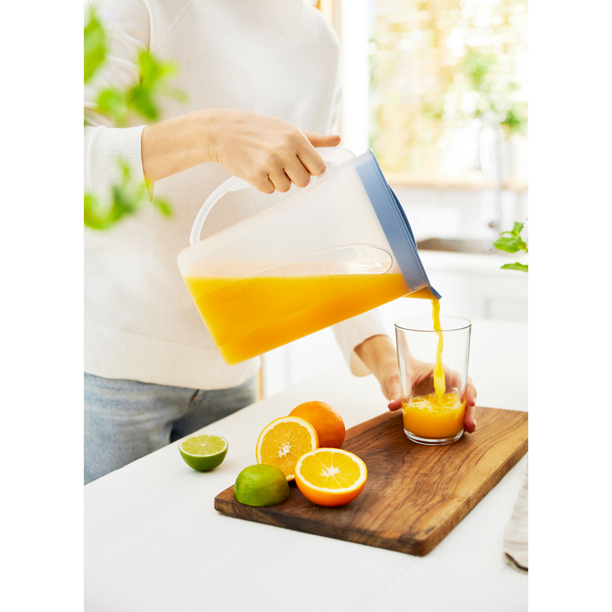 Person gießt ein Getränk aus einer transparenten Kühlschrankkaraffe in ein Glas neben Obst auf einer Arbeitsfläche