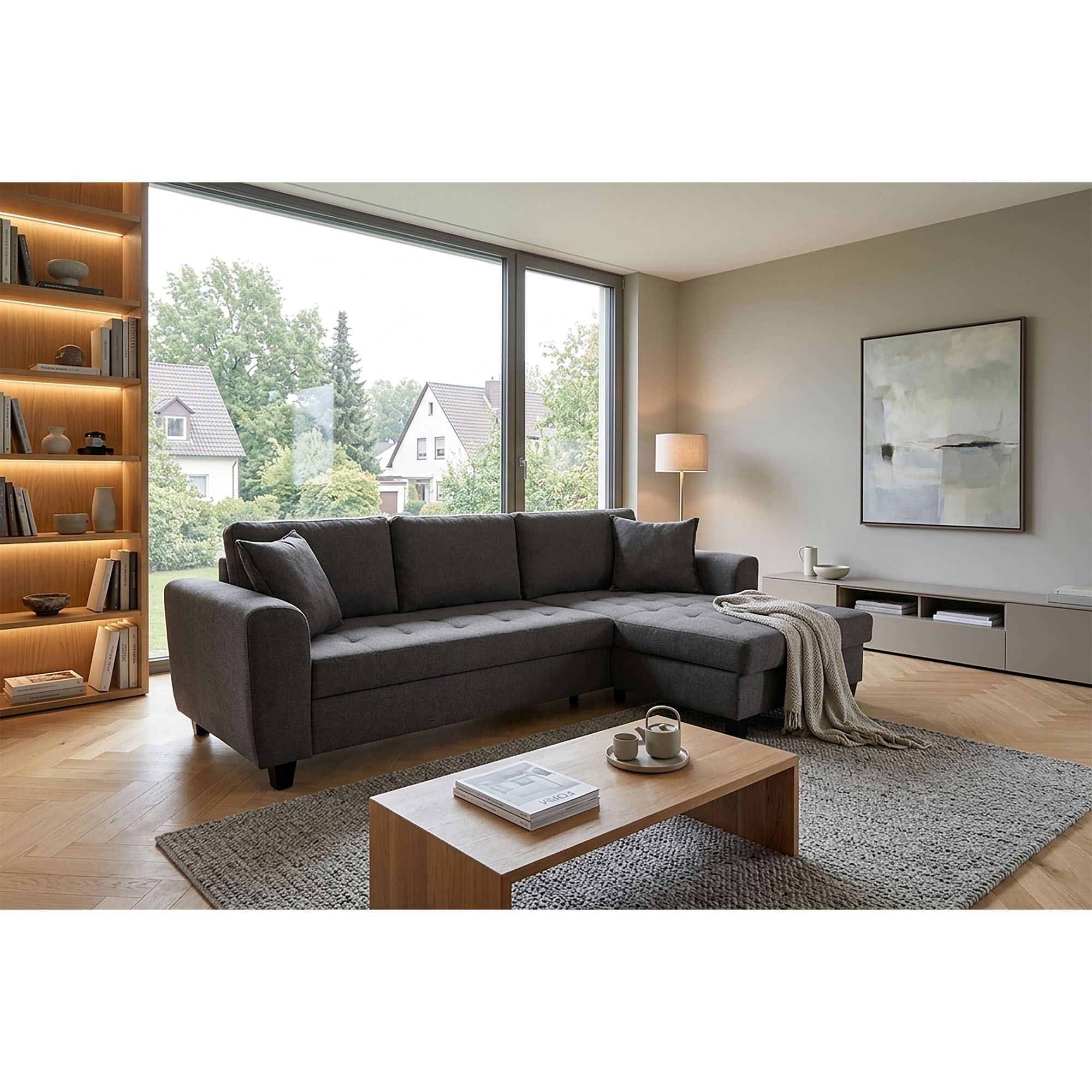 Graues Ecksofa mit rechts angeordneter Ottomane in einem hellen Wohnraum mit großem Fenster, Teppich, Couchtisch und beleuchtetem Bücherregal