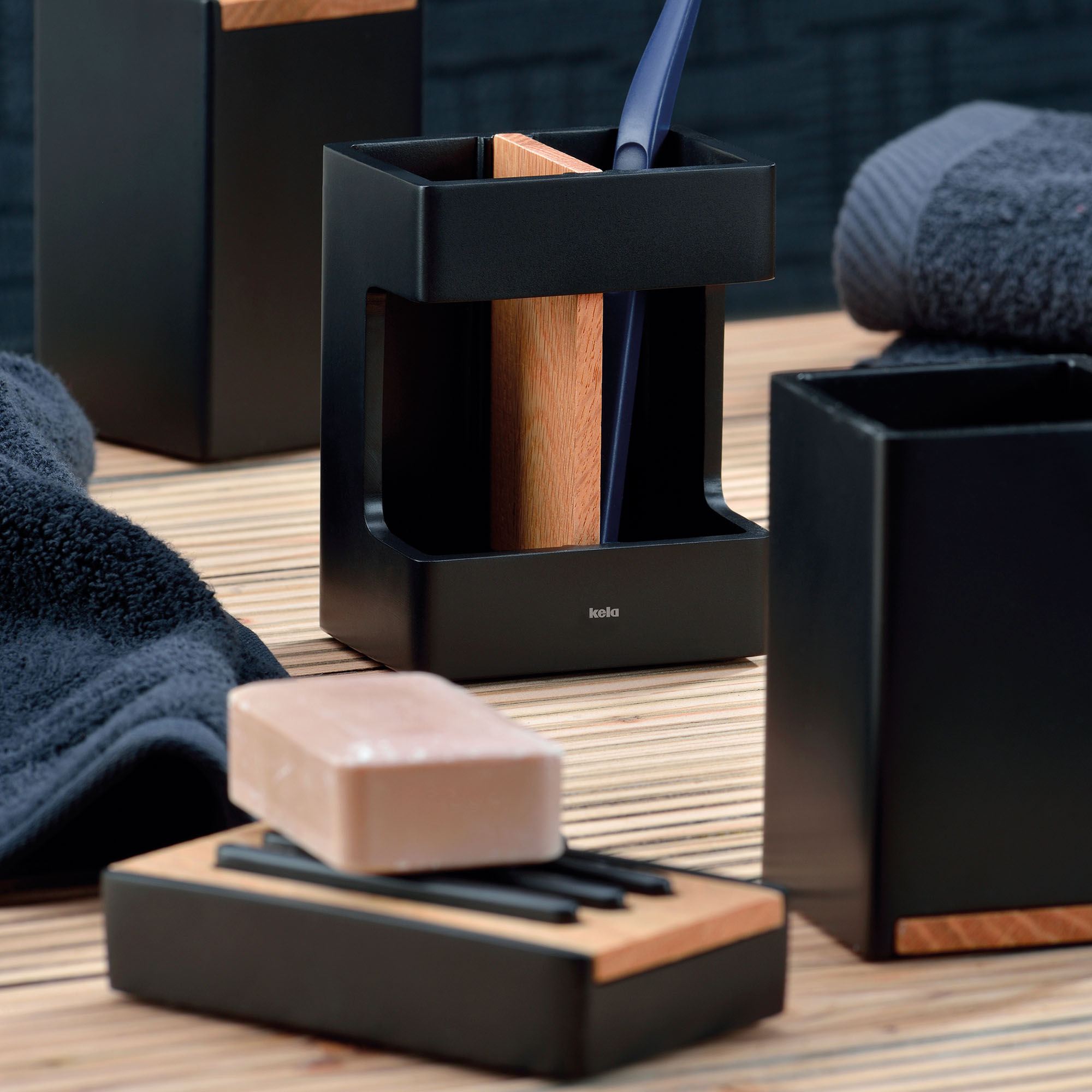 Schwarze rechteckige Seifenschale mit brauner Holzauflage und hellem Seifenstück auf einer Ablage zwischen dunklen Badaccessoires