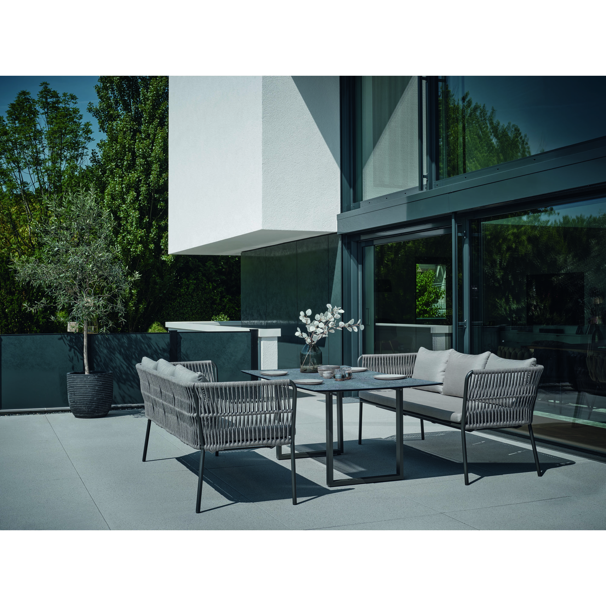 Eine Terrassenszene mit zwei Gartenbänken, einem Tisch und Pflanzen im Hintergrund, vor einer Fassade mit großen Fenstern.