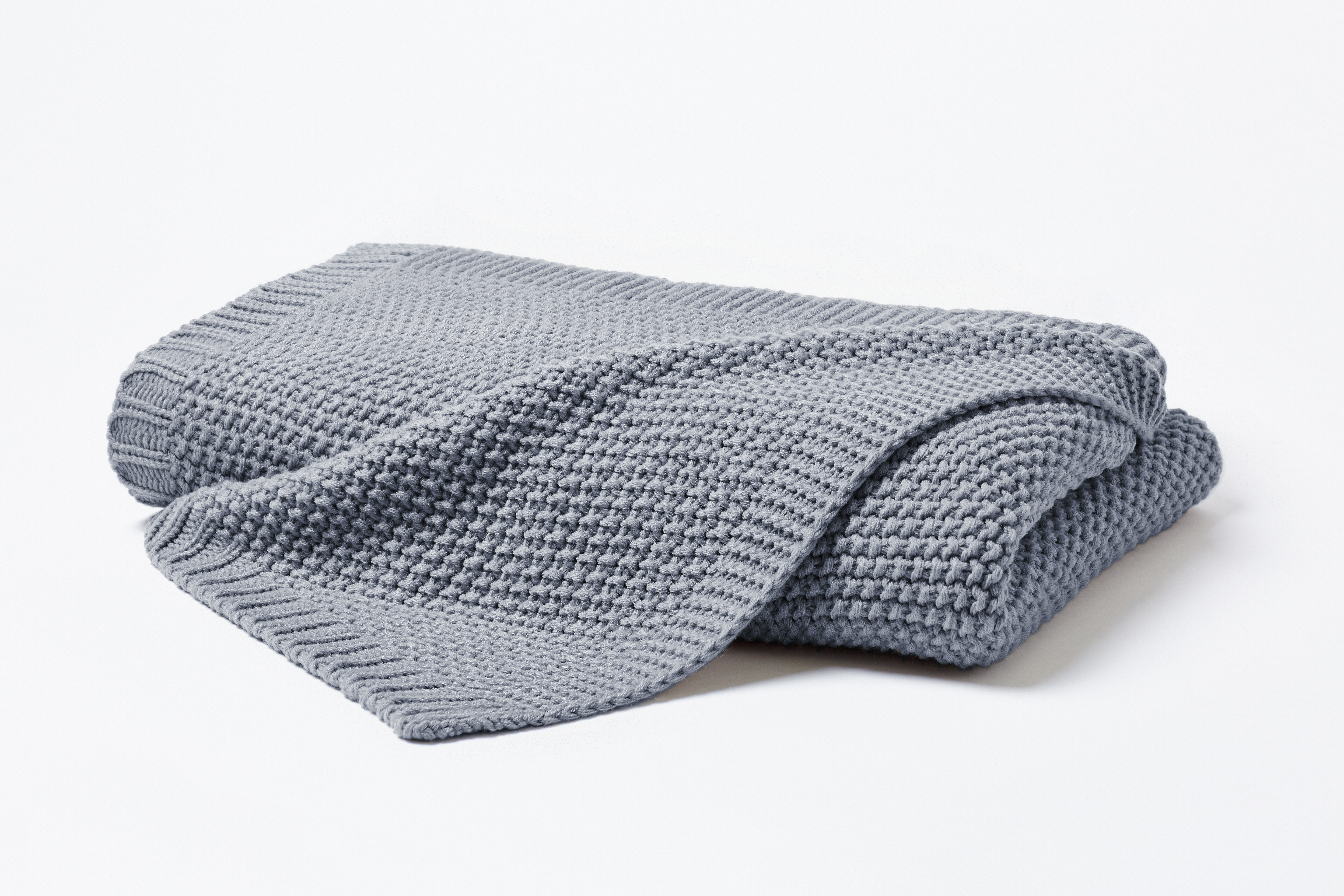Gefaltete Strickdecke in Mittelgrau mit locker übergelegter Ecke; die grobe Waffelstrickstruktur ist vor hellem Hintergrund erkennbar; Größe 130 x 170 cm.