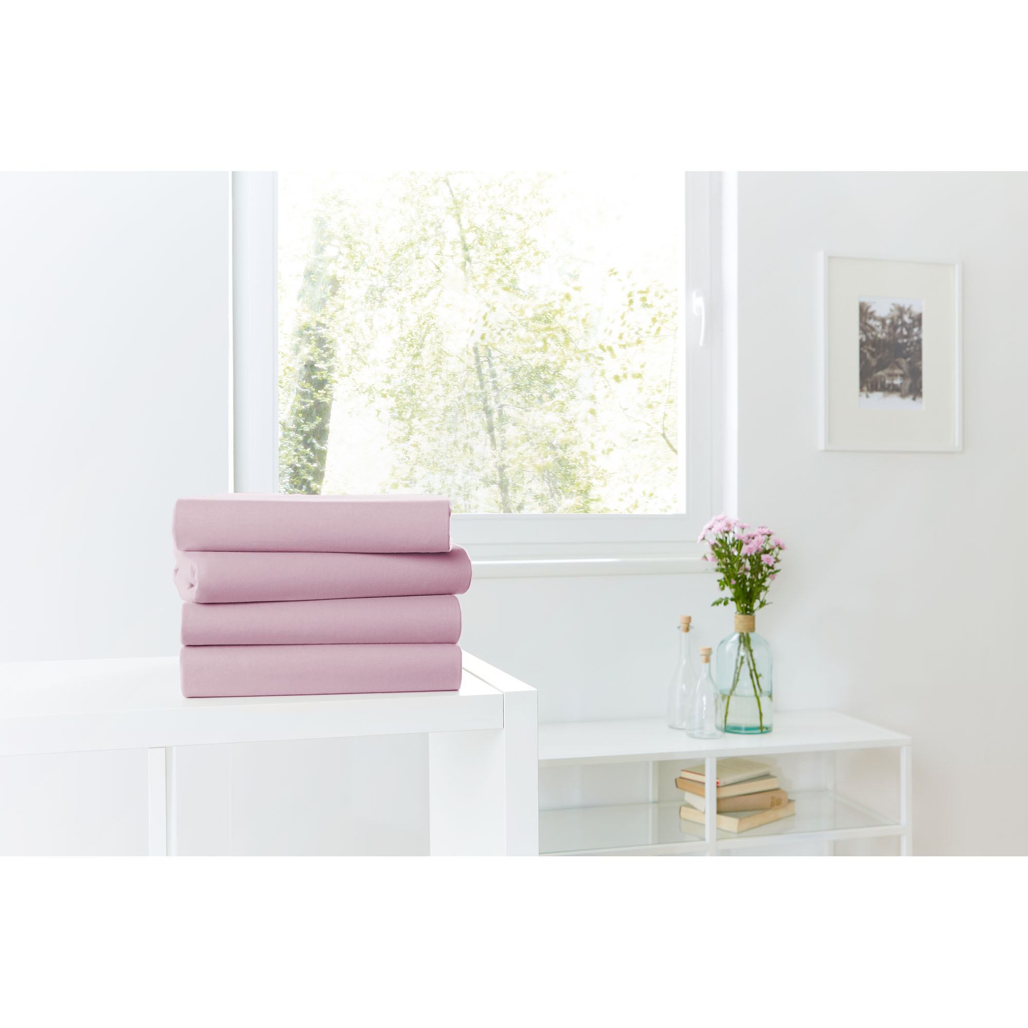 Vier gefaltete Spannbetttücher in Mauve auf einer weißen Kommode vor einem hellen Fenster; Größe 100 x 200 x 30 cm.