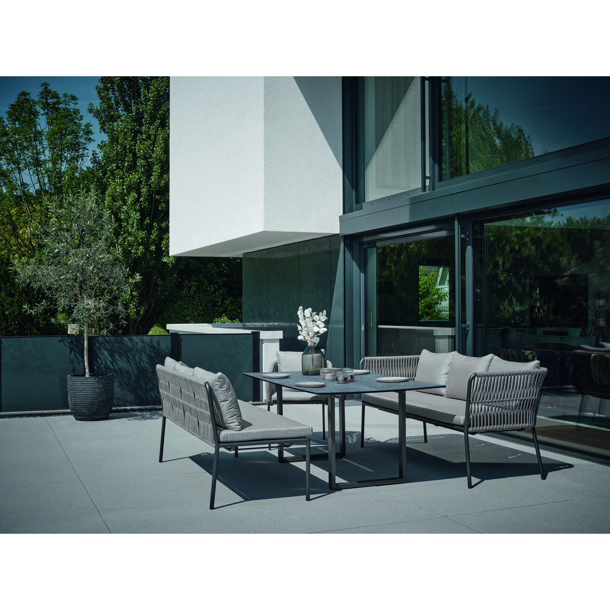 Eine Terrassenszene mit Gartenbänken, Stühlen und einem Tisch vor einem modernen Gebäude mit großen Fenstern und Bäumen im Hintergrund.