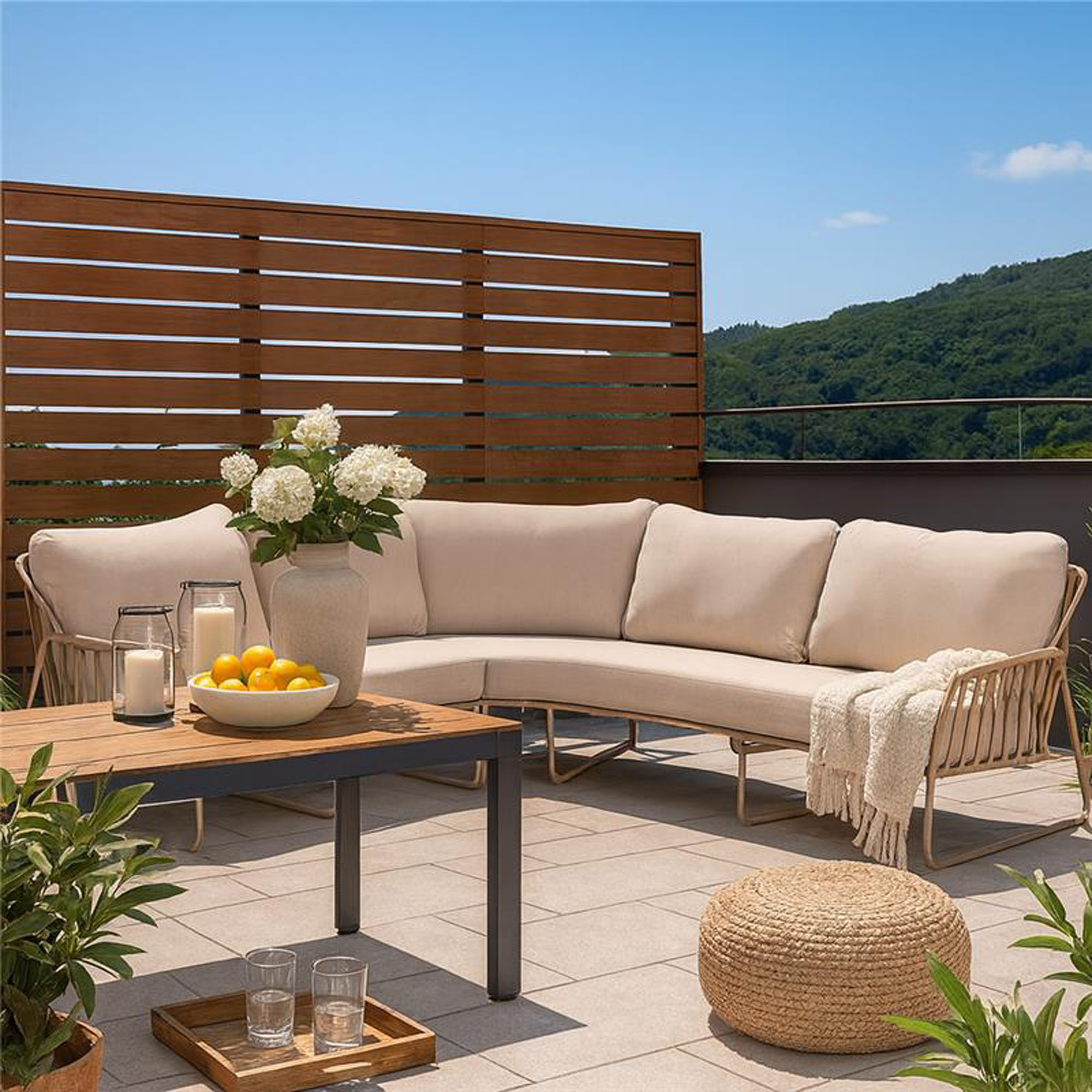 Ecksofa mit beigen Polstern auf einer Terrasse, ergänzt durch Holztisch mit Vase, Zitronenschale und Dekoration, im Hintergrund Sichtschutzwand aus Holz und grüne Landschaft.