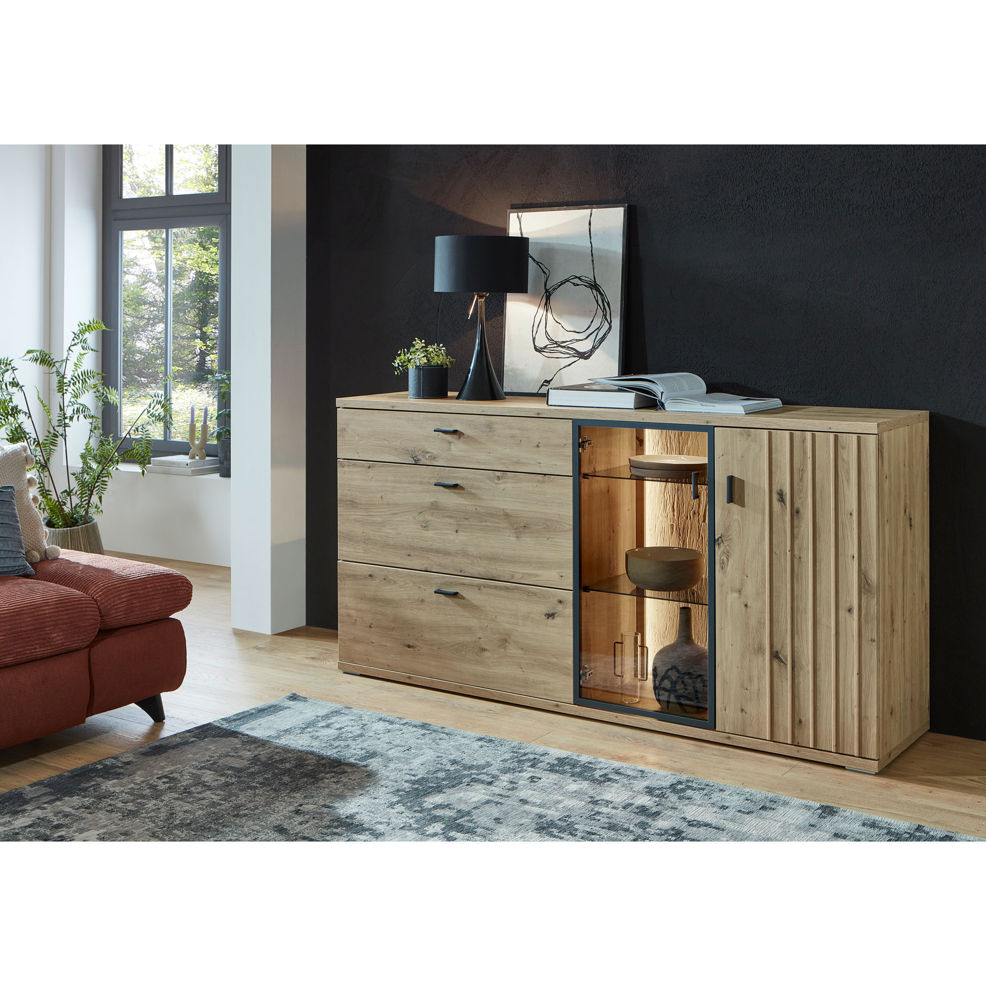 Sideboard in natürlicher Holzoptik mit beleuchtetem Fach und Dekoration vor einer dunklen Wand im Wohnraum