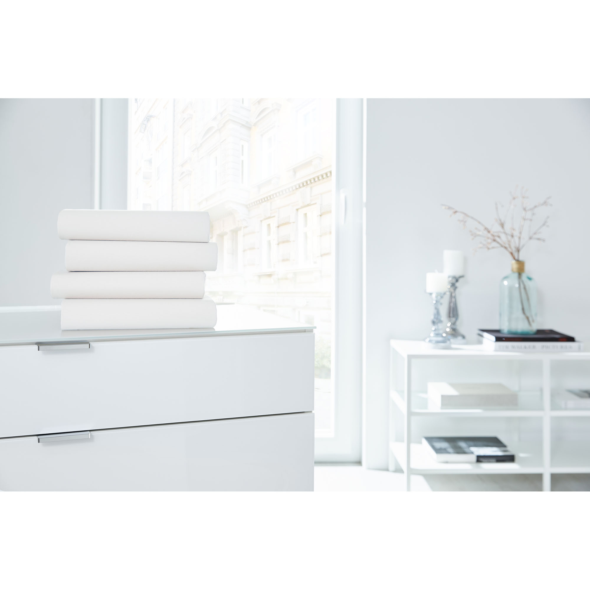 Vier gefaltete Boxspring-Spannbetttücher in Weiß auf einer weißen Kommode in einem hellen Raum; Größe 100 x 200 x 38 cm.