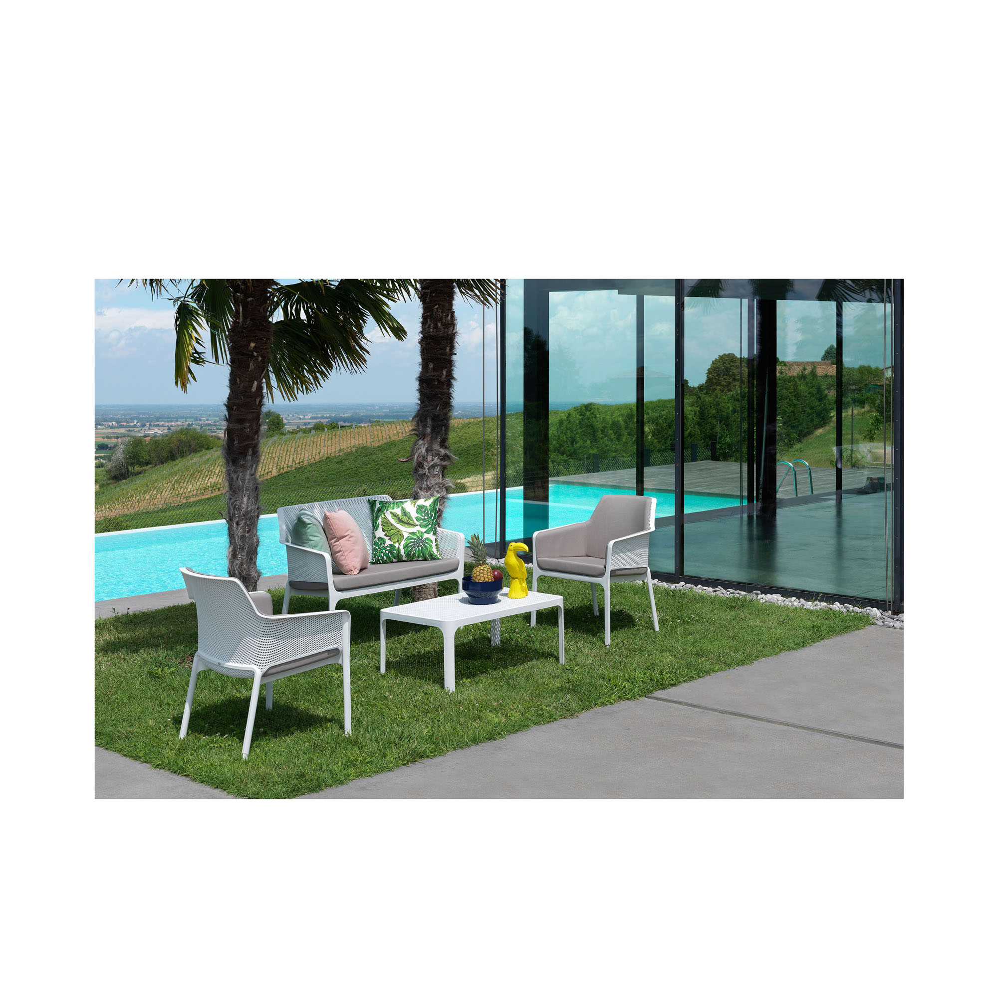 Eine moderne Outdoor-Sitzgruppe steht auf einer Rasenfläche neben einem Pool. Sie besteht aus weißen Stühlen und einer weißen Bank mit grauen Sitzkissen, dazu einem weißen Tisch mit Dekoobjekten. Im Hintergrund sind Palmen, Glasfassade und hügelige Landsc