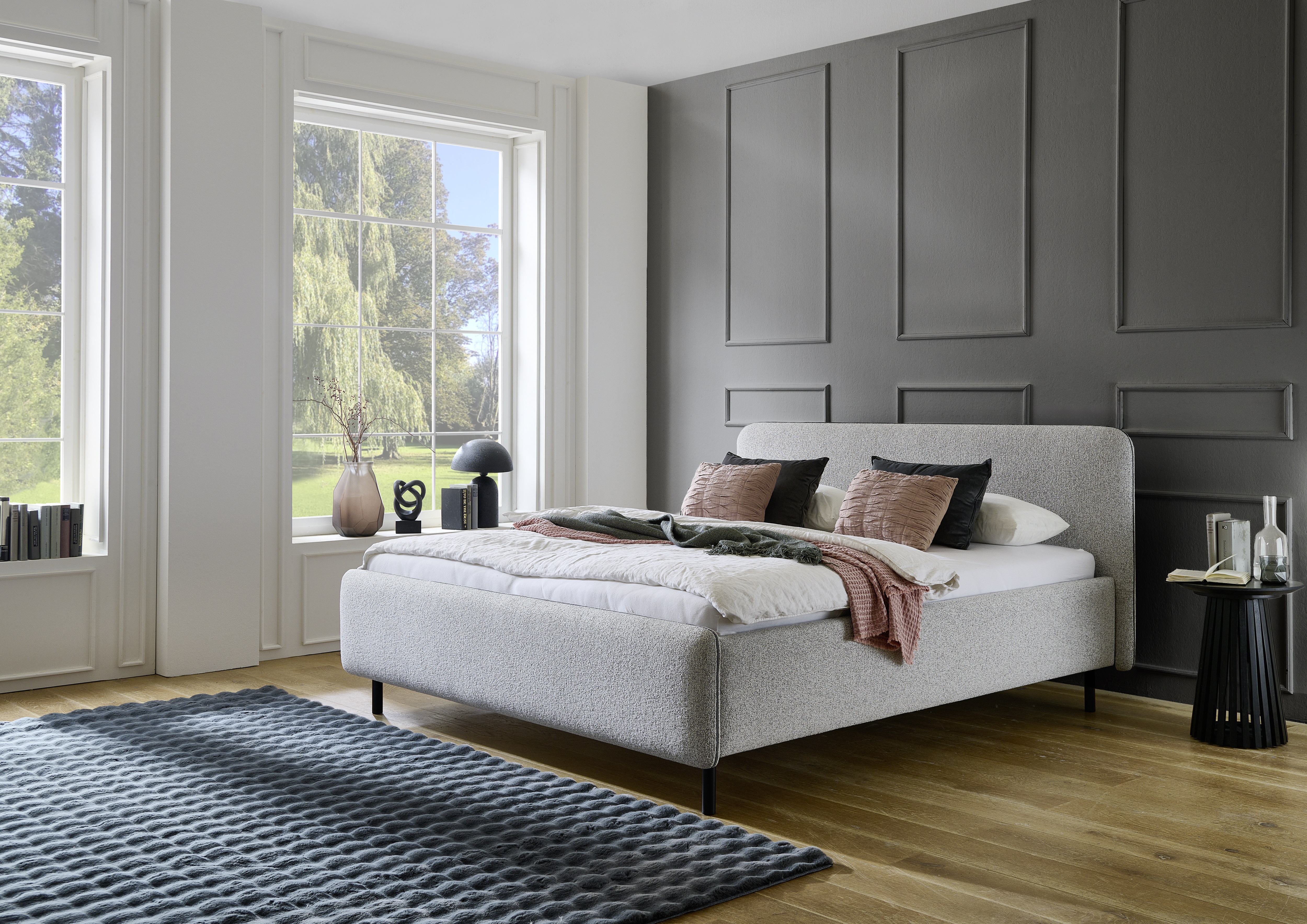 Helles Polsterbett mit rundem Kopfteil in einem modernen Schlafzimmer mit dunkler Akzentwand, großen Fenstern und blauem Teppich auf Holzboden