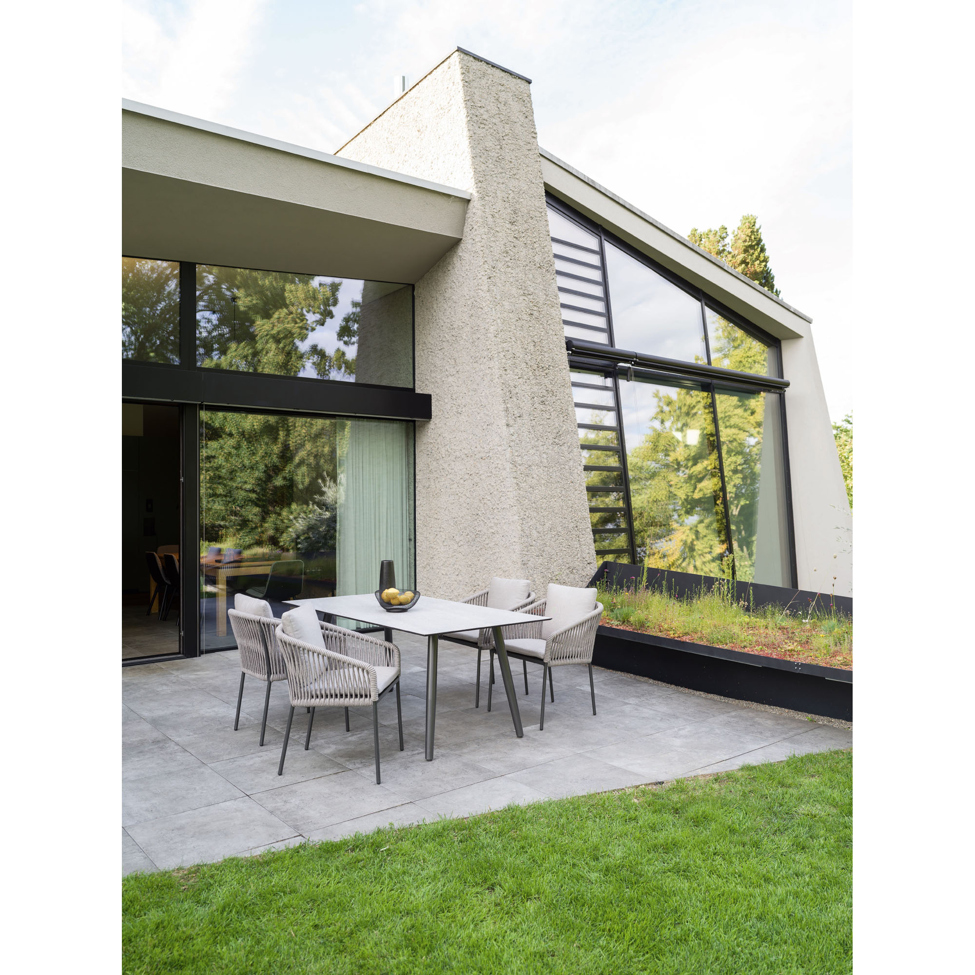 Eine moderne Terrasse mit vier beigen Stühlen aus Seilgeflecht und einem rechteckigen Tisch mit heller Platte. Die Terrasse liegt vor einem Haus mit großen Glasfronten und Blick auf grüne Bäume.