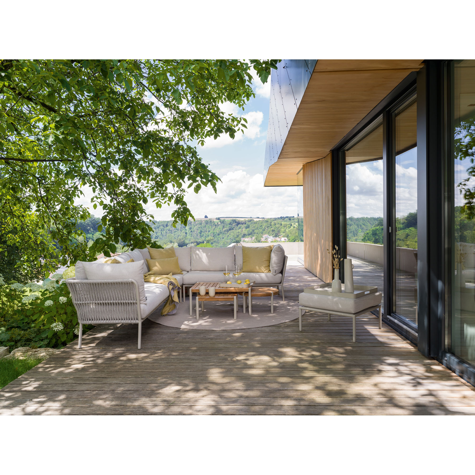 Moderne Terrasse mit hellen Lounge-Möbeln und einem niedrigen Holztisch, umgeben von Bäumen und mit Blick auf eine grüne Landschaft.