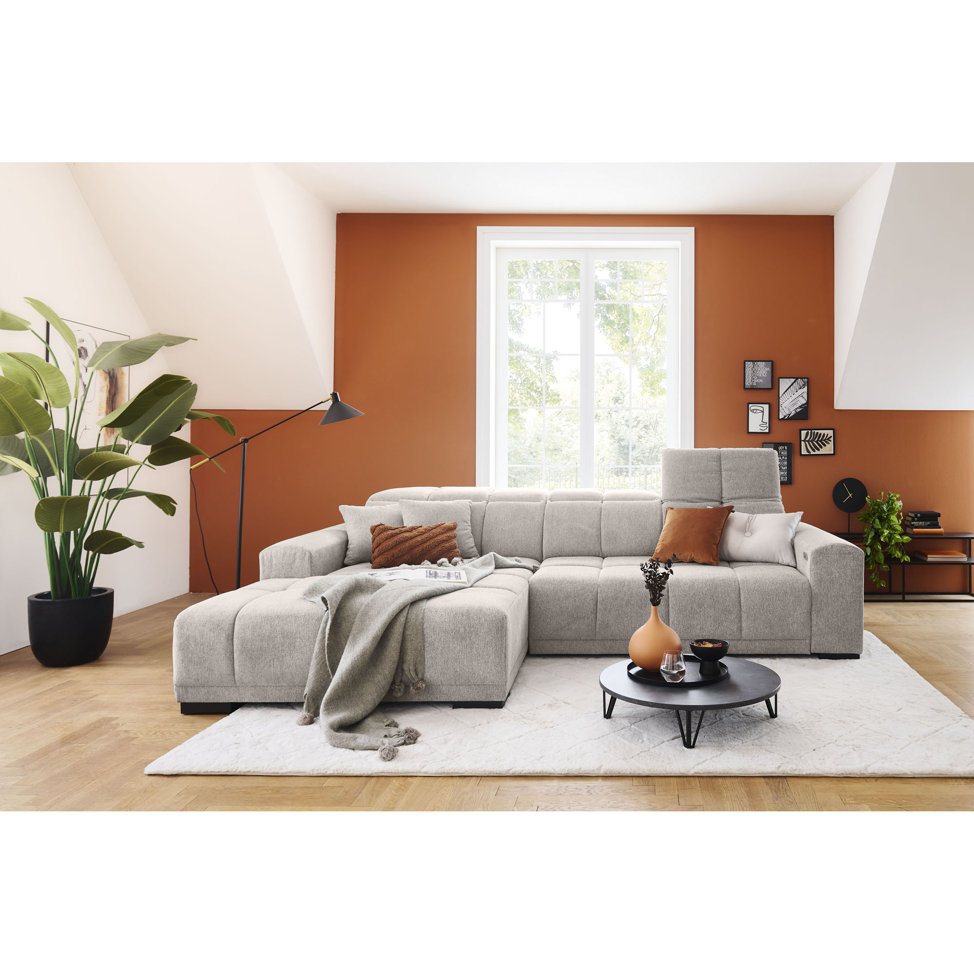 Silbergraue Wohnlandschaft mit Ottomane links im Bild, verstellbarer Kopfstütze, mehreren Kissen und Decke in einem Wohnzimmer mit orangefarbener Wand