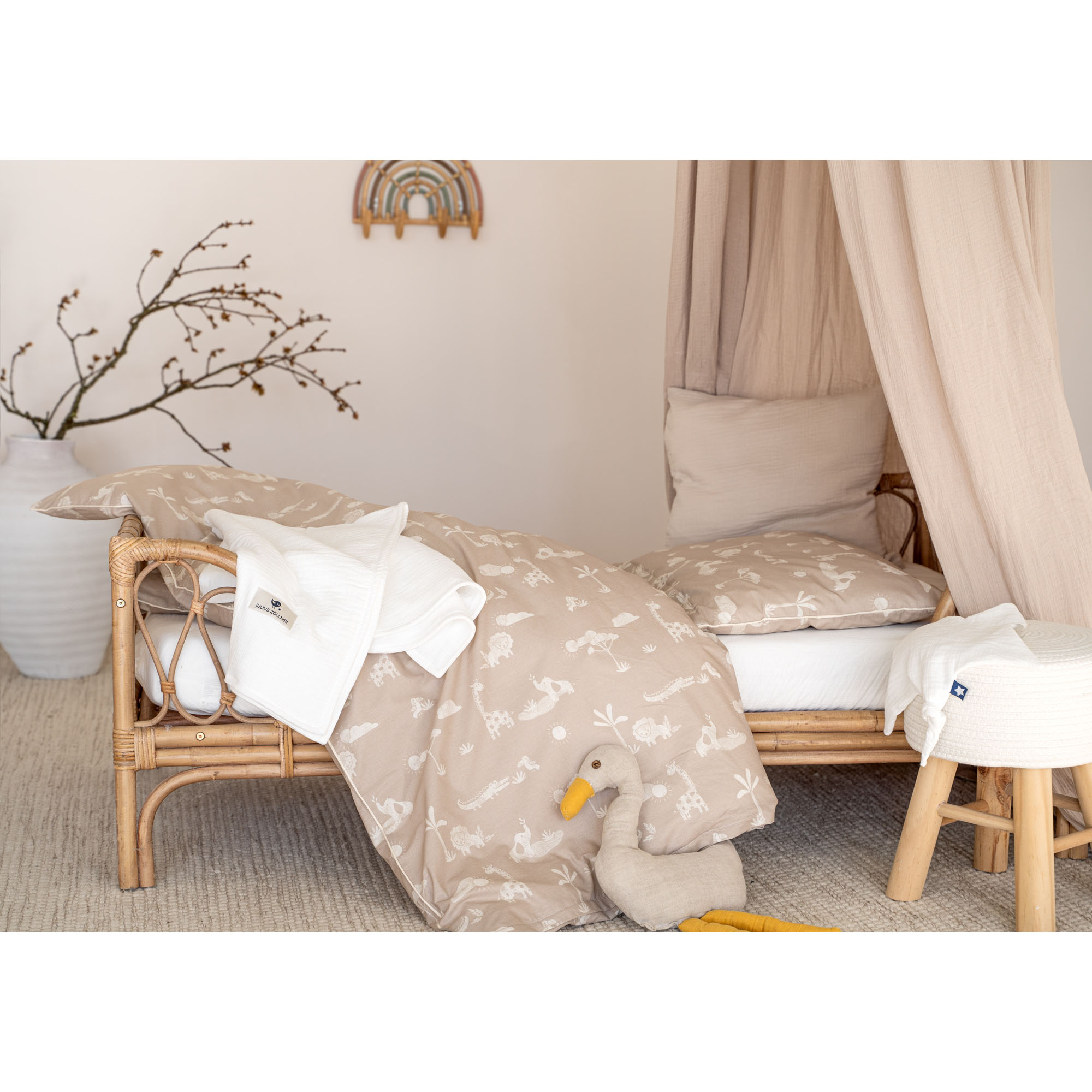 Beigefarbene Kinderbettwäsche mit weißen Safari-Motiven in einem Rattanbett mit Baldachin, Kuscheltier und heller Raumdekoration