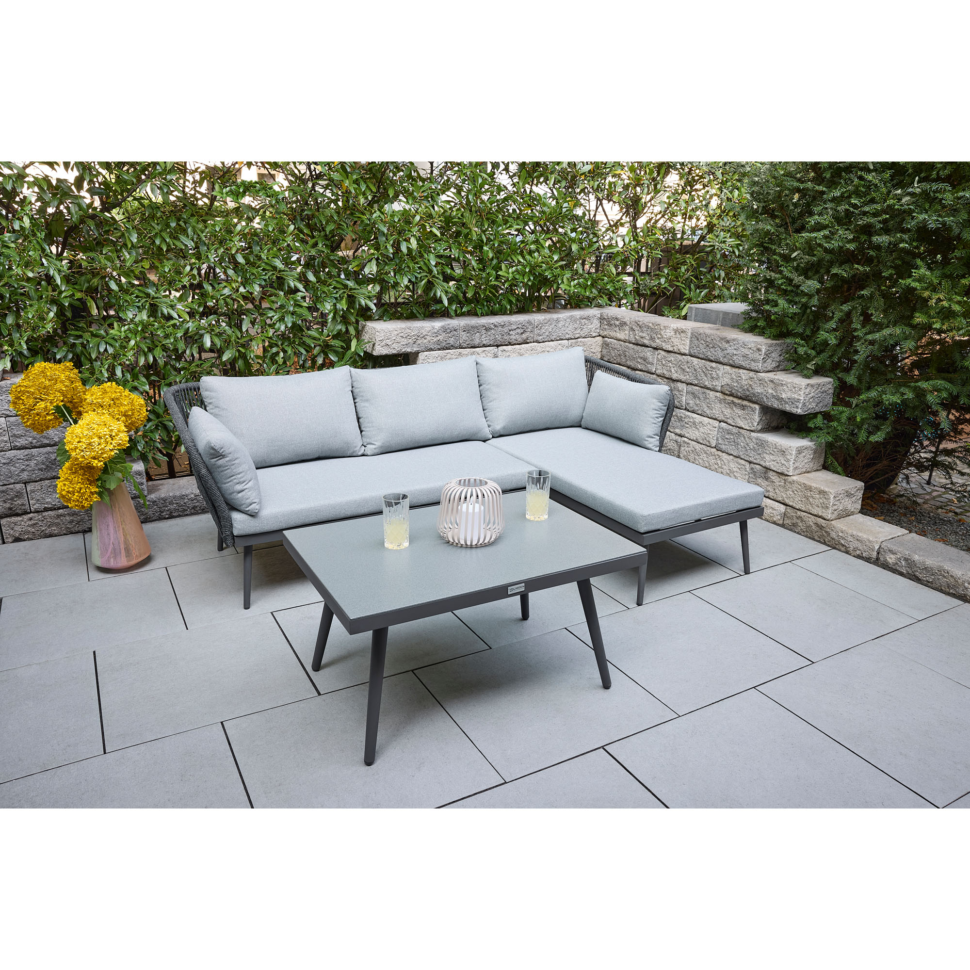 Gartenlounge mit Ecksofa und Tisch auf einer Terrasse im Garten mit Hecke und Steinmauer im Hintergrund