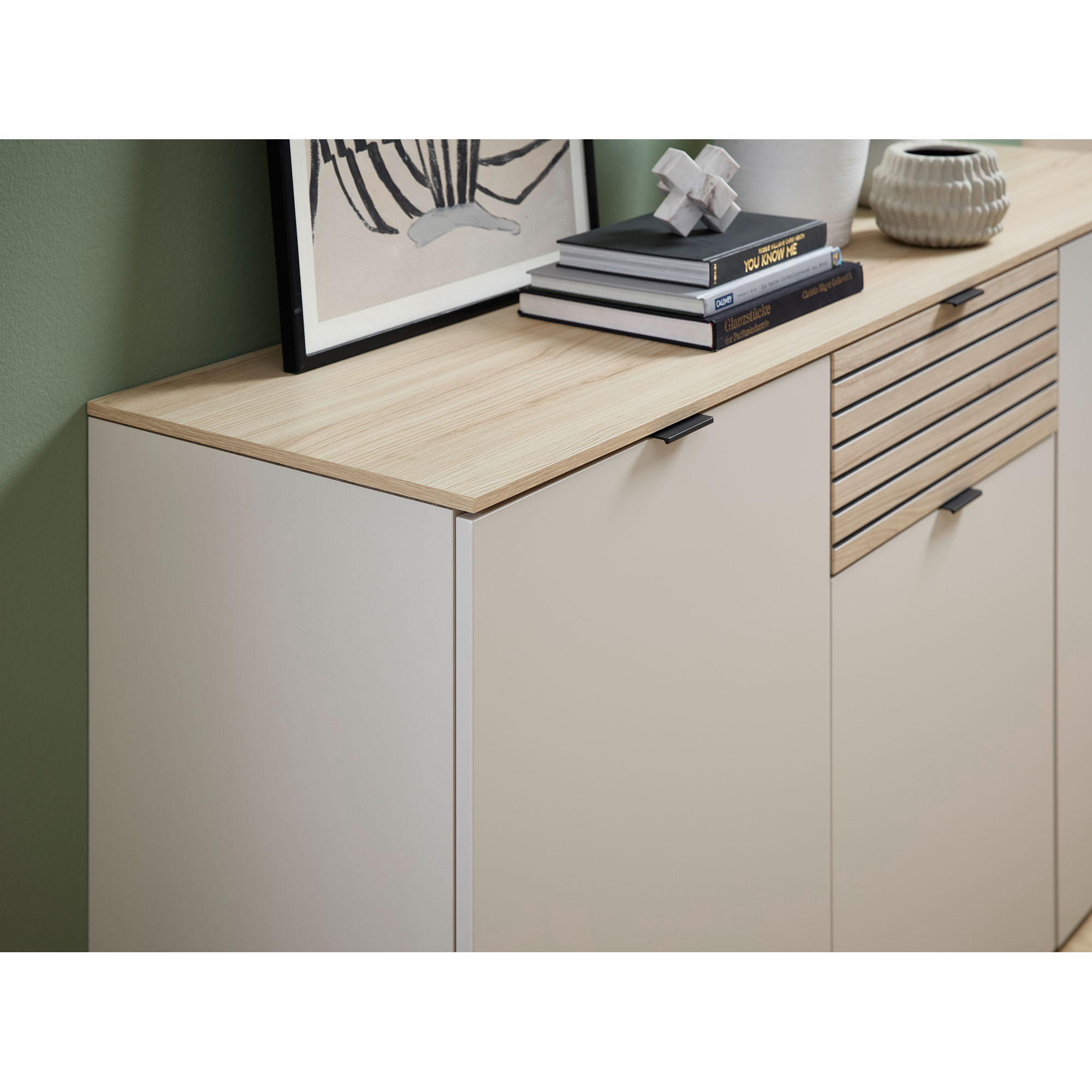 Nahaufnahme des Sideboards mit heller Holzoberplatte und schlichten Fronten. Ein Teil der Dekoration und der Bücher ist sichtbar.