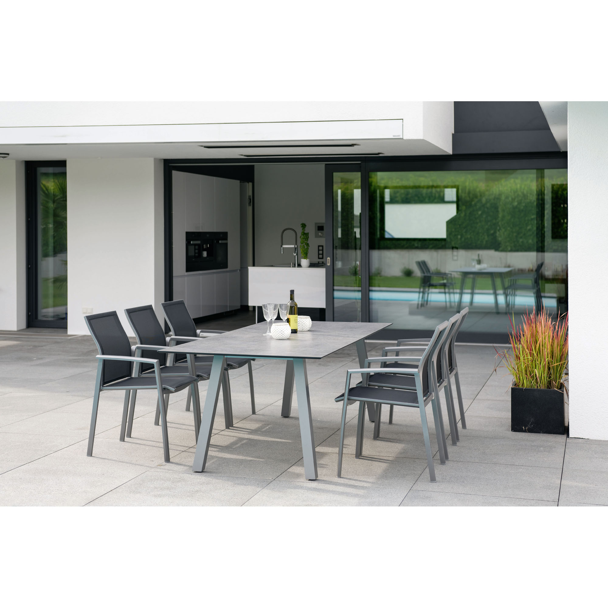 Mehrere graue Gartenstühle stehen um einen rechteckigen Tisch auf einer modernen Terrasse mit Glasfassade und Pflanzen.