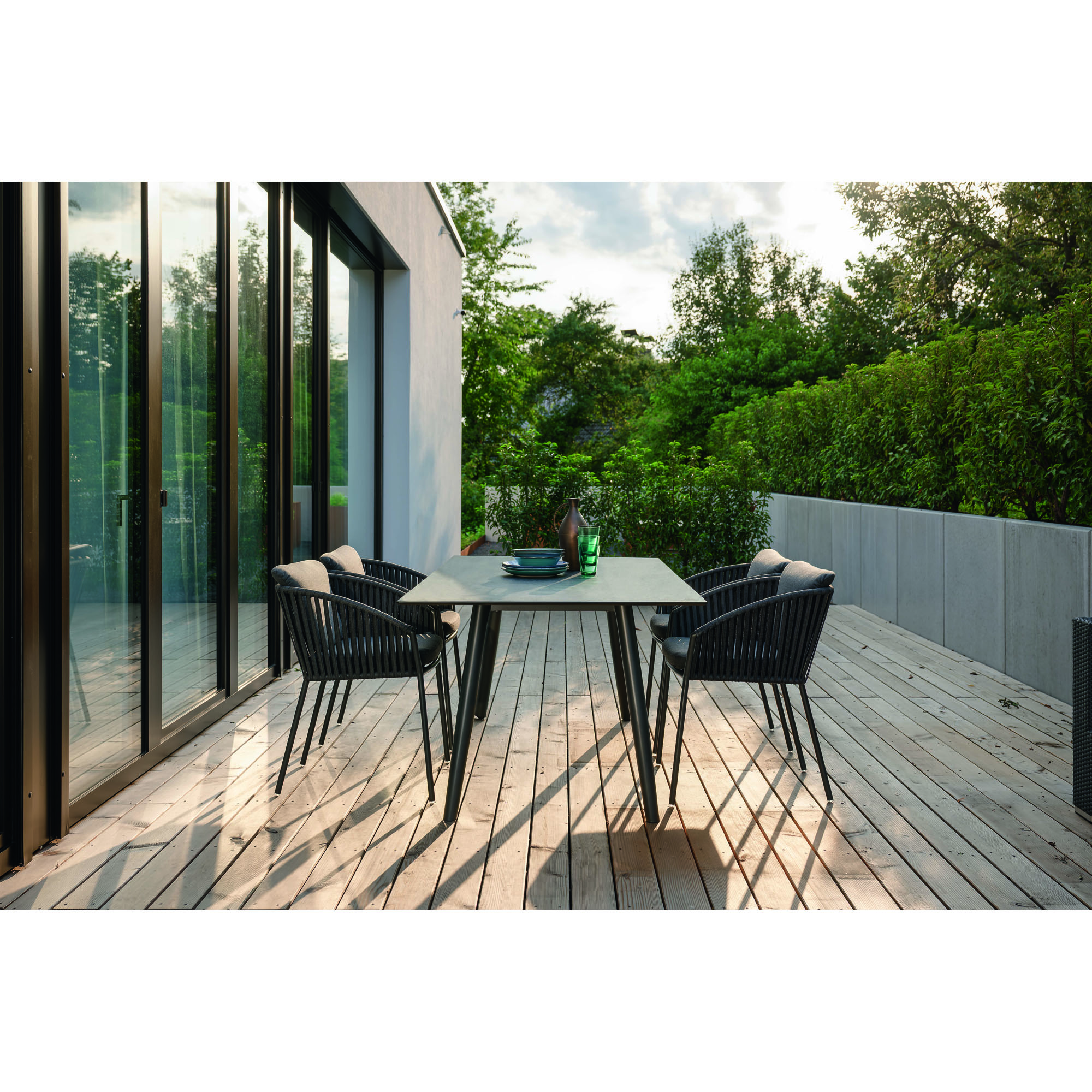 Rechteckiger Tisch mit vier Stühlen auf einer Holzterrasse neben einem Gebäude mit Glasfront