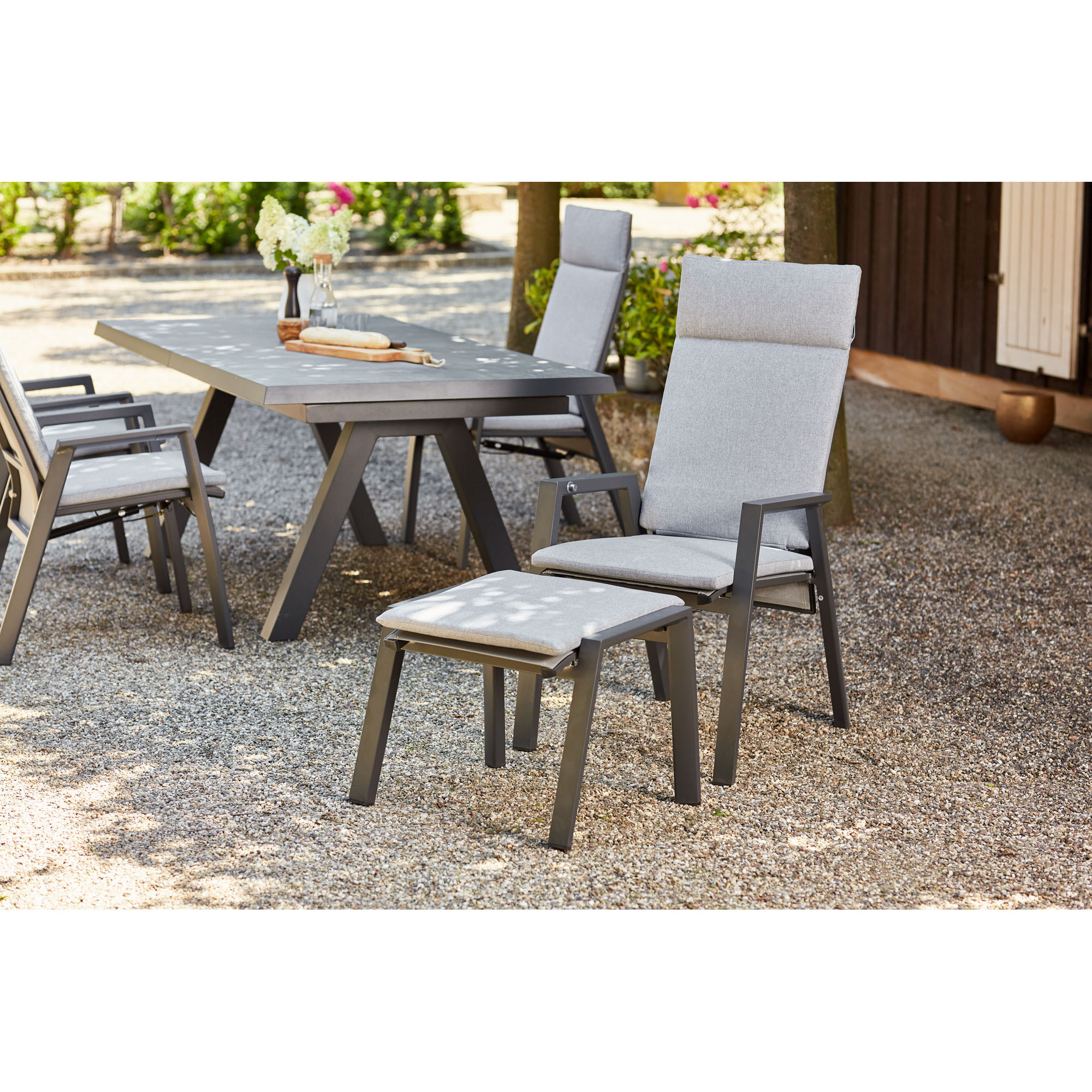 Ein Hocker mit grauem Polster steht vor einem Stuhl an einem großen Gartentisch, umgeben von weiteren Stühlen auf einem Kiesboden.
