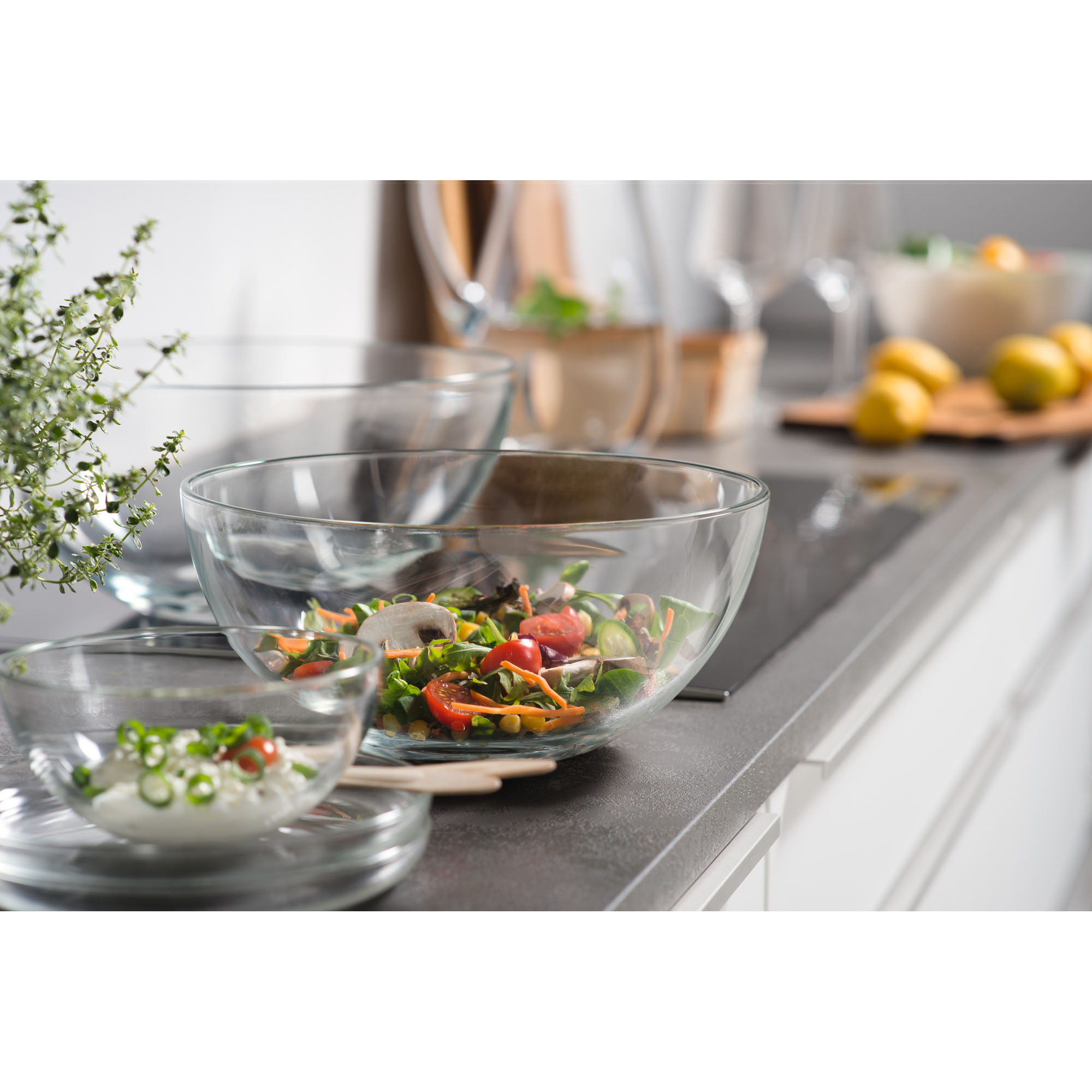 Mehrere Glasschalen mit Salat und Zutaten auf Küchenarbeitsplatte mit Pflanzen und Gemüse im Hintergrund