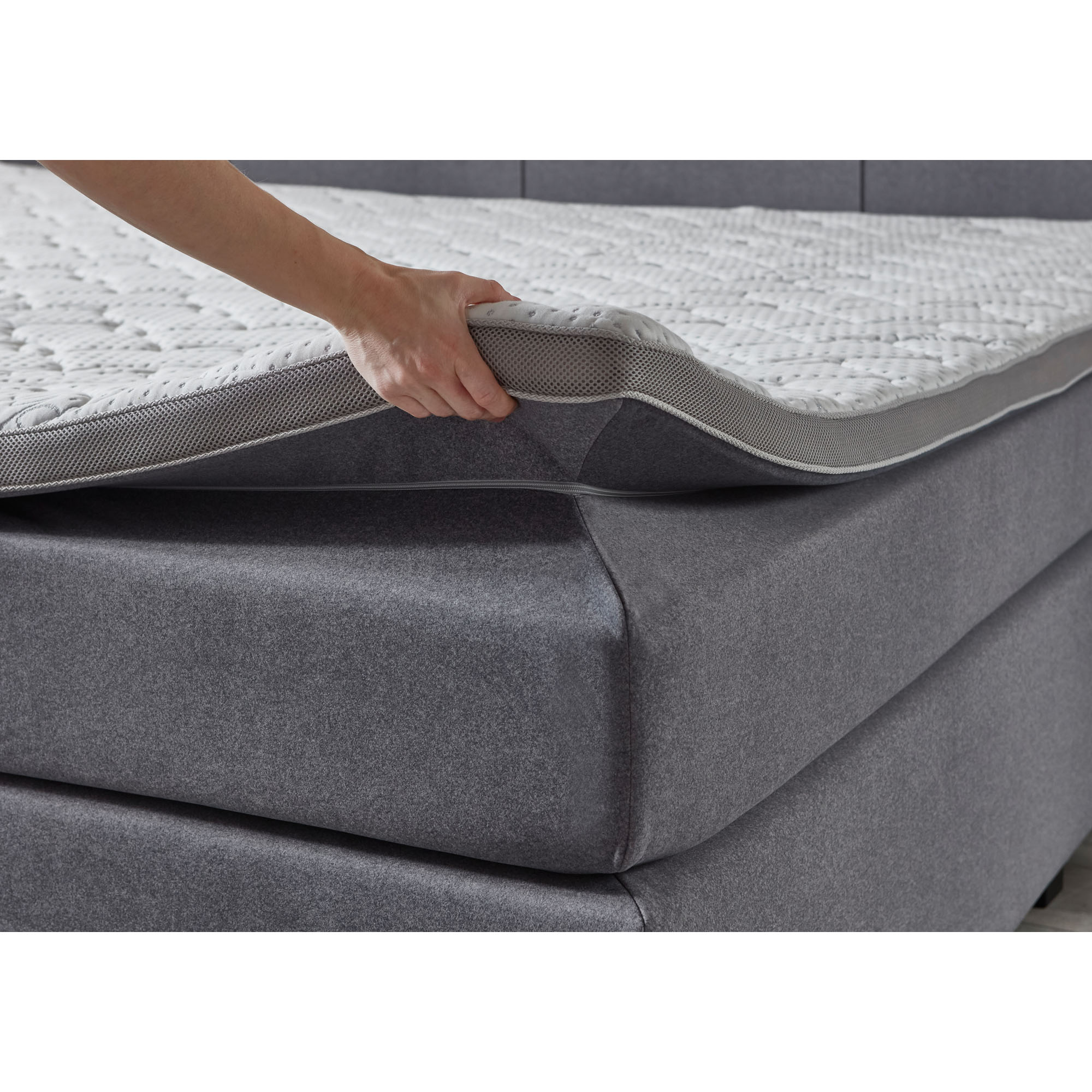 Boxspringbett mit grauem Stoffbezug während ein Topper an der Ecke angehoben wird