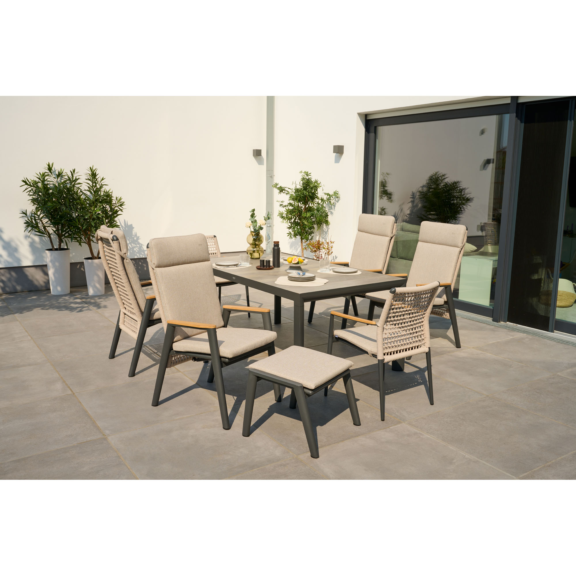 Stilvolles Gartenmöbel-Set mit Tisch, Stühlen und Hocker auf sonniger Terrasse, umgeben von Pflanzen und Glasfront.