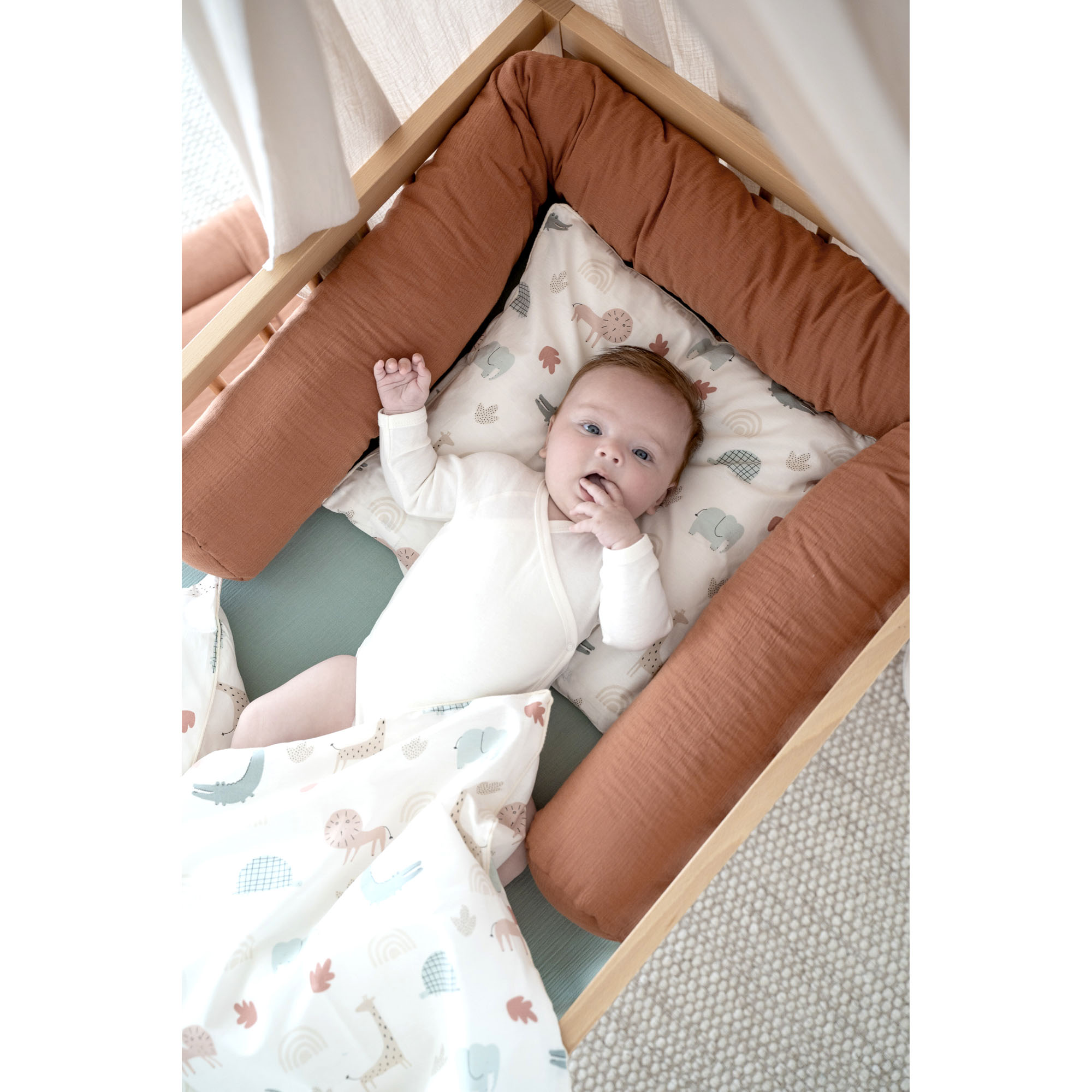 Baby liegt in einem Holzbett mit terracottafarbener Musselin-Bettschlange und bunter Tierbettwäsche