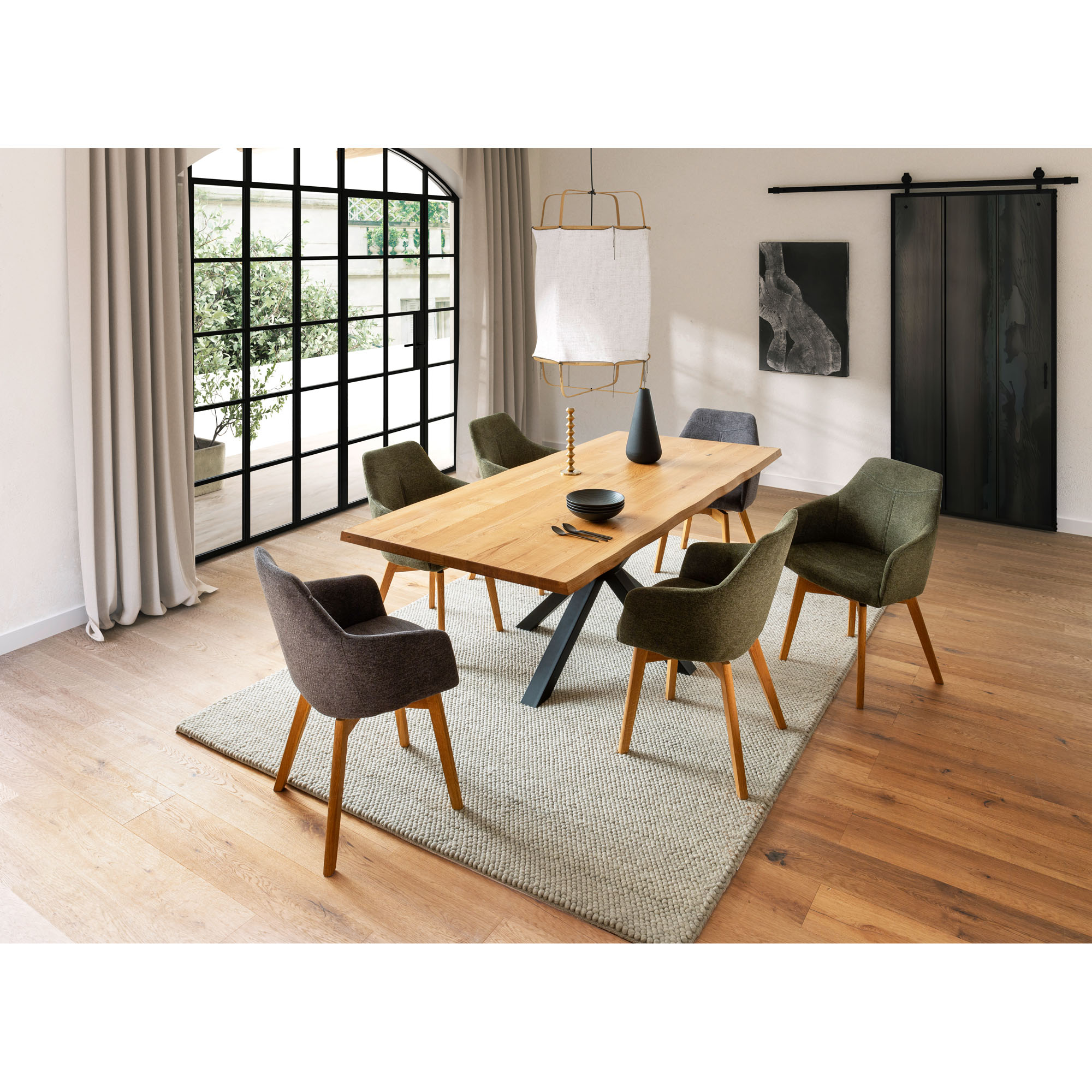Holz-Esstisch mit schwarzem Metallgestell, umgeben von sechs Polsterstühlen in verschiedenen Grüntönen und Braun. Der Tisch steht auf einem Teppich in einem hellen Raum mit großen Fenstern und Schiebetür.