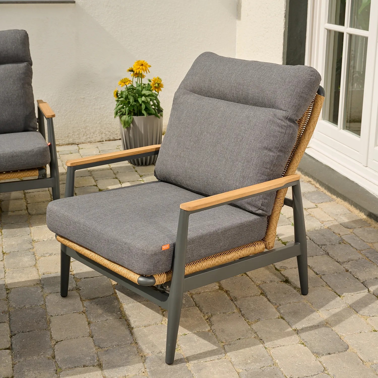 Ein Sessel mit dunklem Gestell, seitlich sichtbarem Geflecht und grauen Sitz- und Rückenpolstern, auf einer Terrasse platziert.