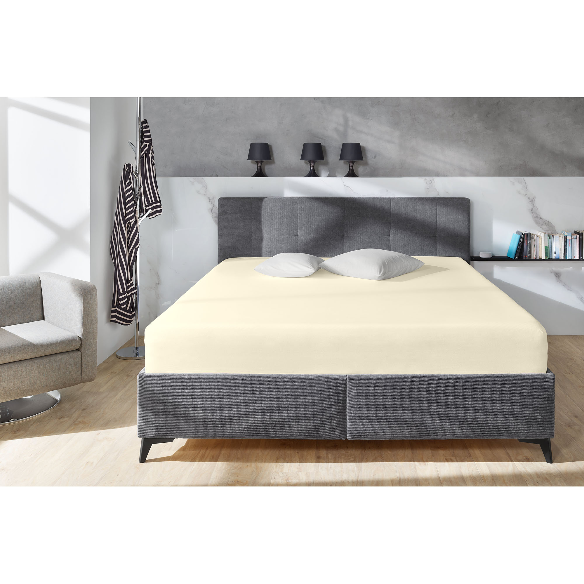 Boxspring-Spannbetttuch in Ecru auf einem grauen Boxspringbett in einem Schlafzimmer; zwei helle Kissen liegen am Kopfende; Größe 100 x 200 x 38 cm.